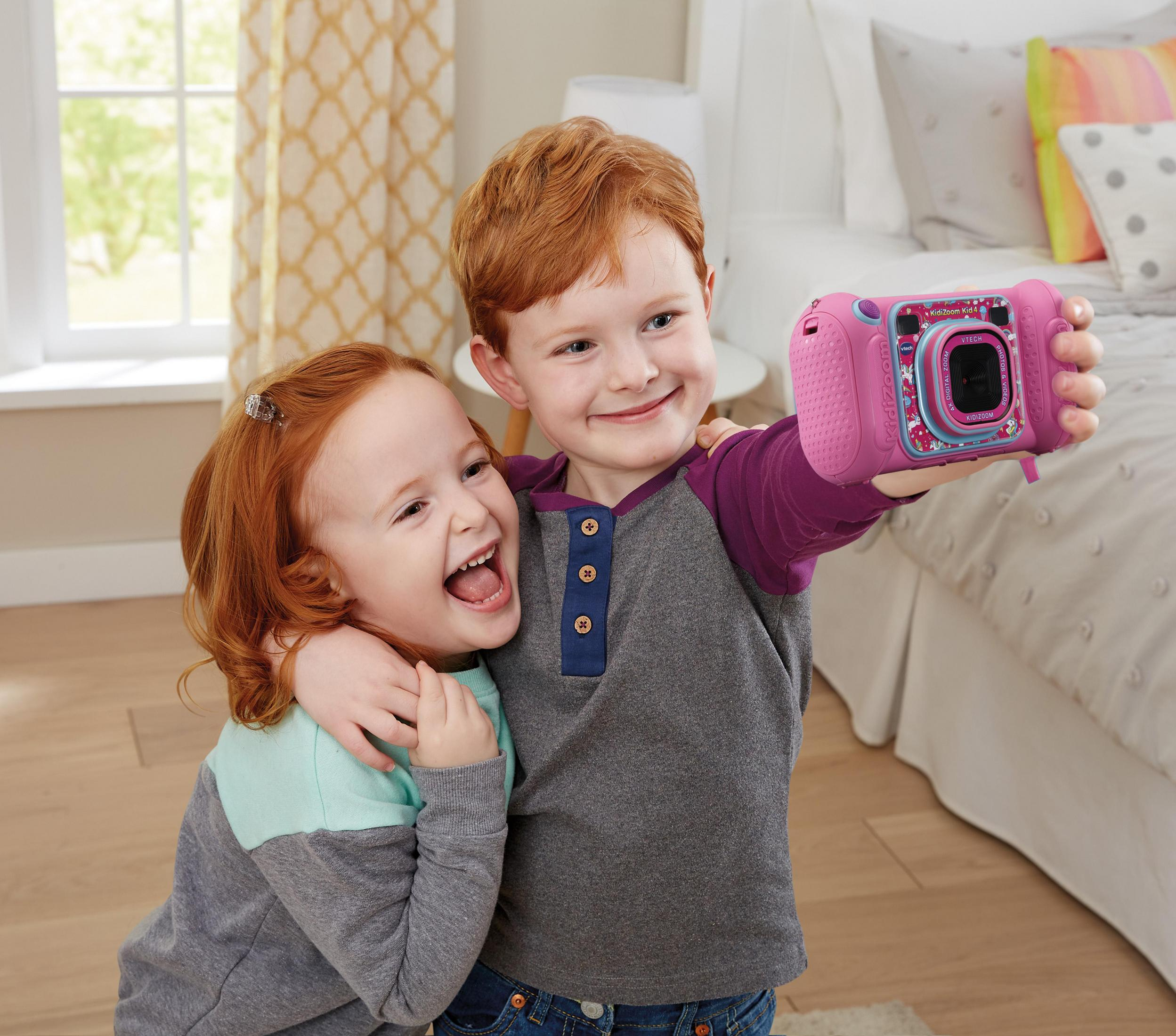 Zwei Kinder machen ein Selfie mit einer pinken Kamera, lächeln und umarmen sich in einem Raum.