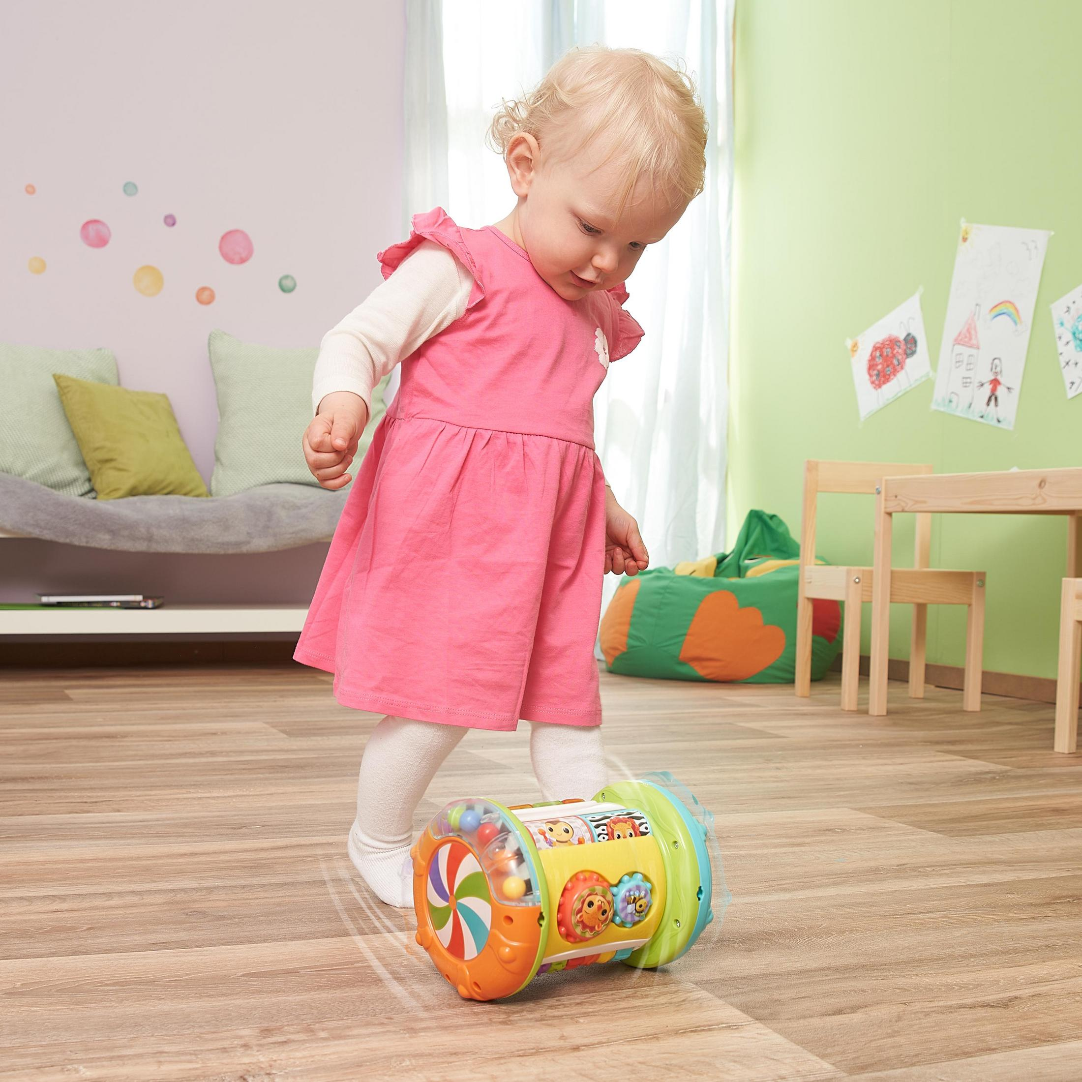 Kleines Mädchen in rosa Kleid spielt mit rollendem Spielzeug mit bunten Elementen auf Holzboden.