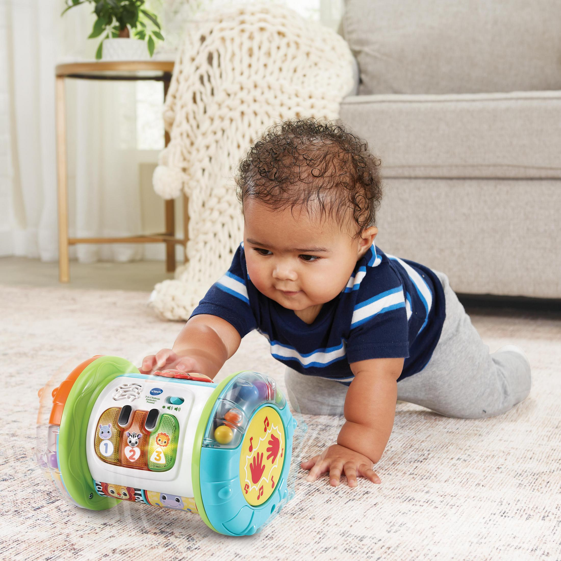 Ein Baby krabbelt auf eine bunte Spielzeugtrommel zu. Die Trommel hat Knöpfe, Lichter und einen Griff.