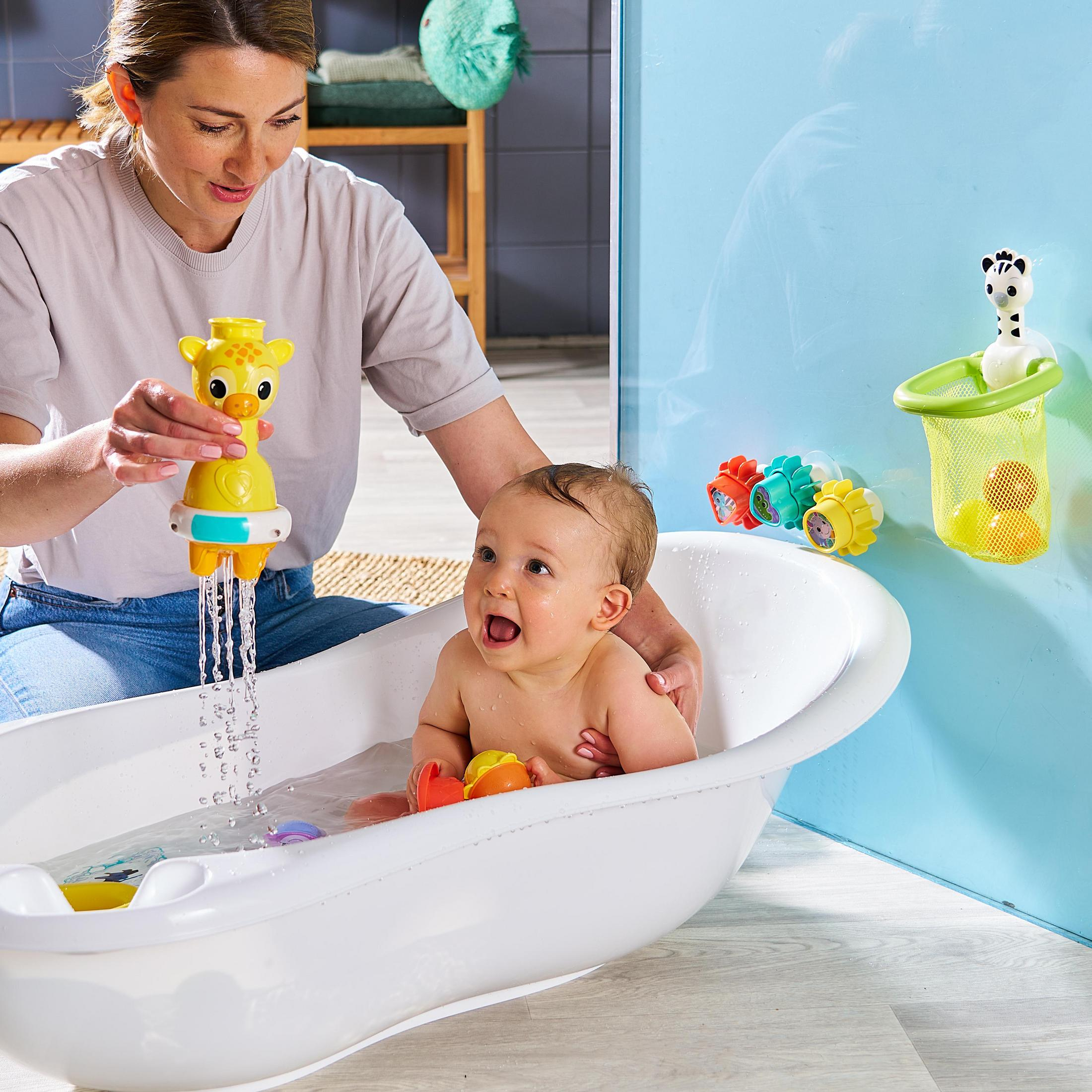 Baby in der Badewanne wird von einem Erwachsenen gehalten. Ein Giraffenspielzeug sprüht Wasser. Ein Zebraspielzeug in einem Netz.