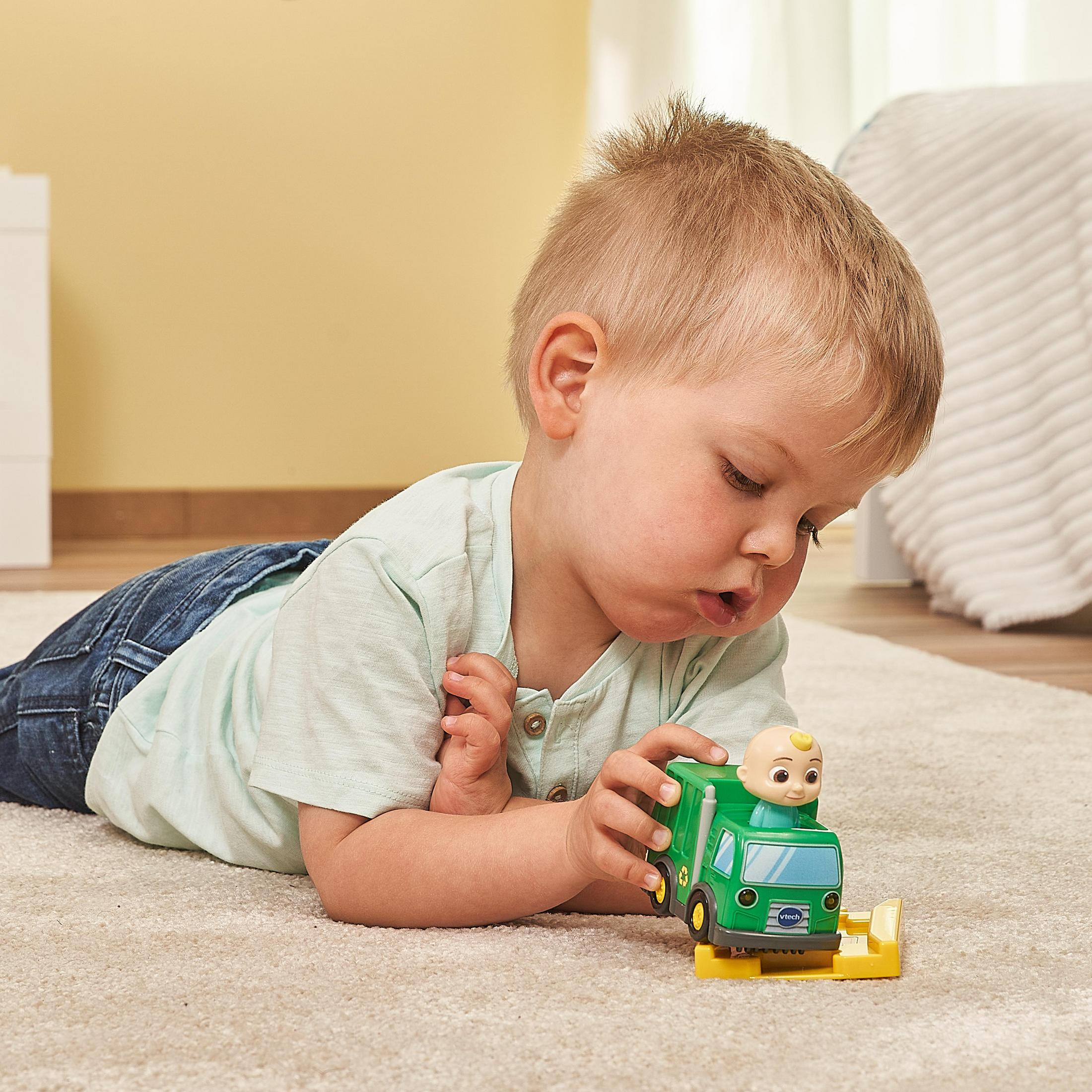 Ein Kleinkind liegt auf einem Teppich und betrachtet einen grünen Spielzeug-LKW mit einer Babyfigur darauf.