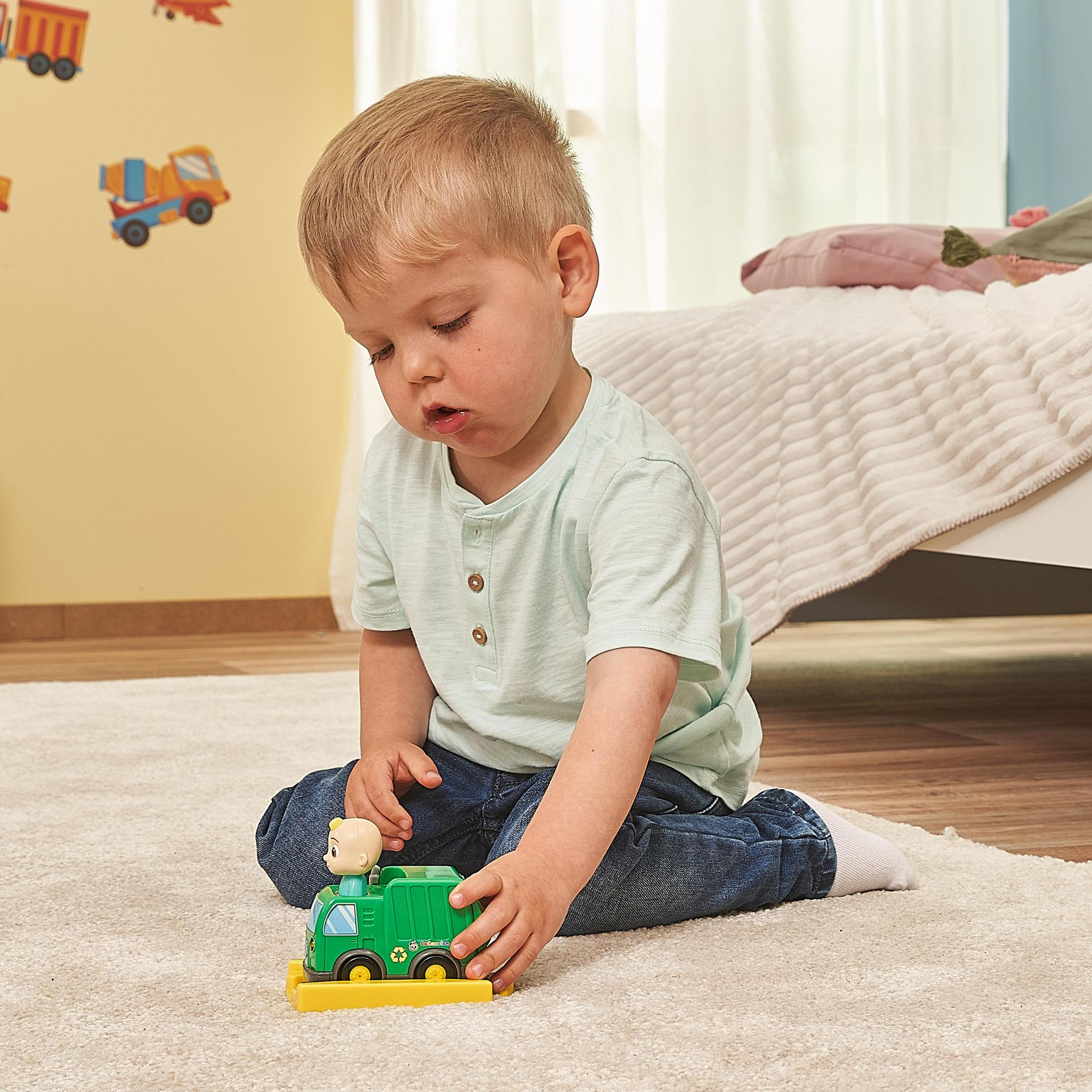 Junge spielt mit einem grünen Müllwagen-Spielzeug, sitzt auf einem Teppich, trägt hellblaues Hemd und Jeans.