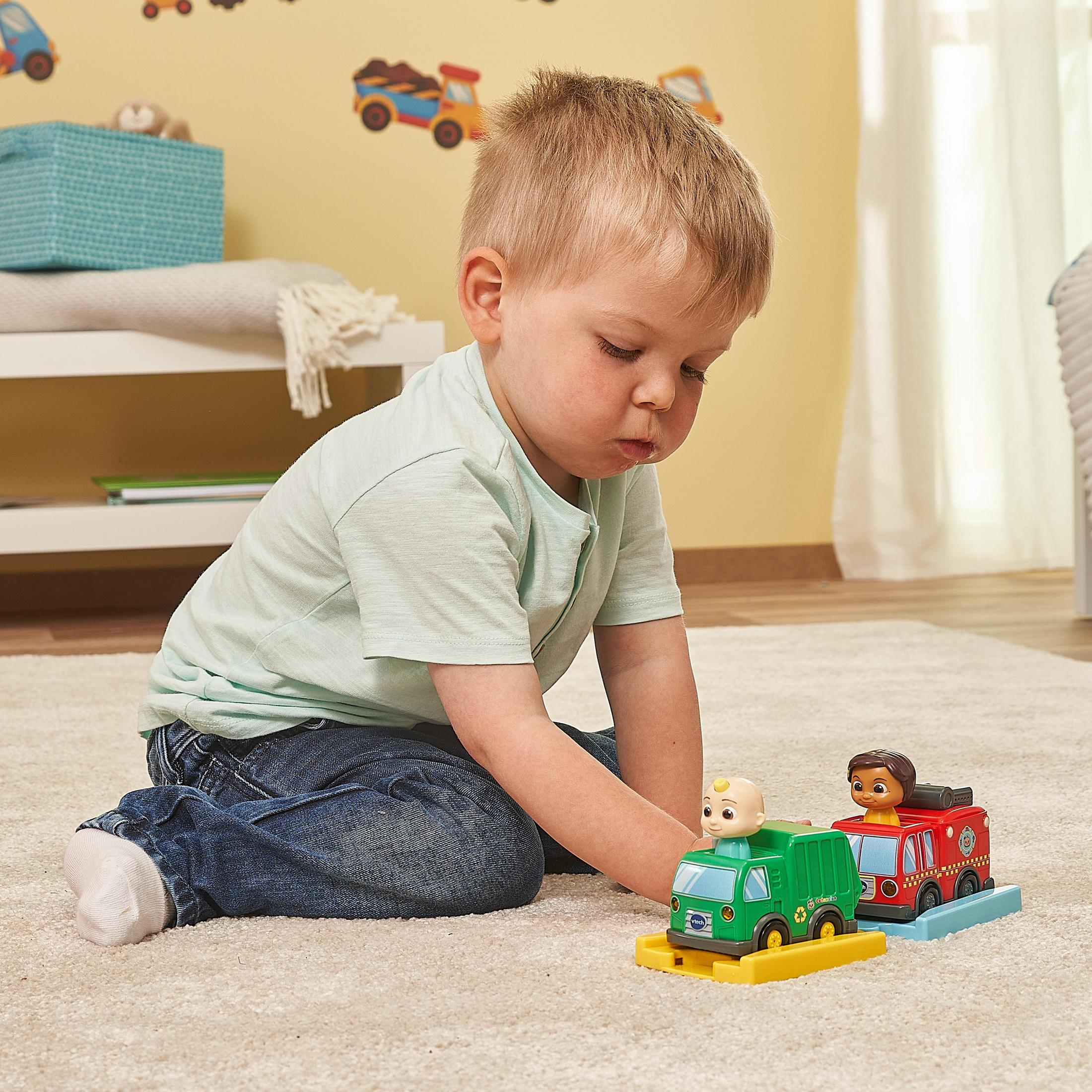 Ein Kind spielt mit Spielzeug-LKWs auf einem Teppich. Ein weiterer Spielzeug-LKW und ein Feuerwehrauto sind vorhanden.