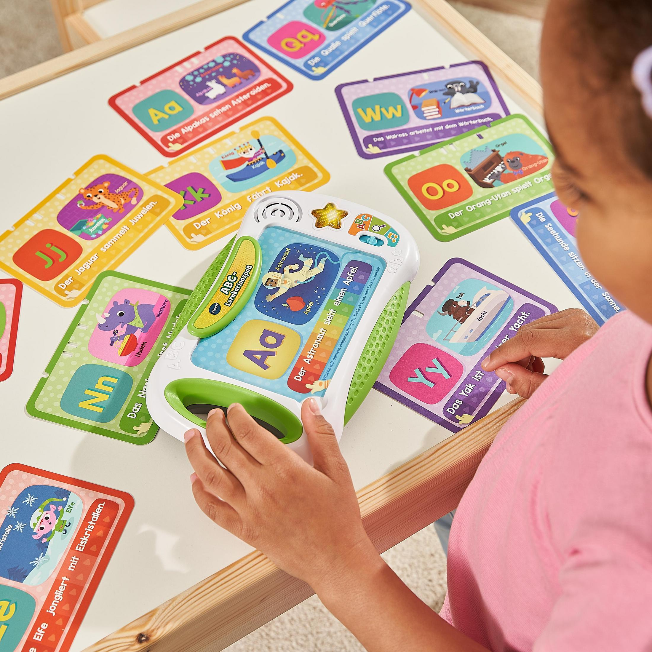 Ein Mädchen hält ein Spielzeug-Tablet, umgeben von Karteikarten auf einem weißen Tisch.
