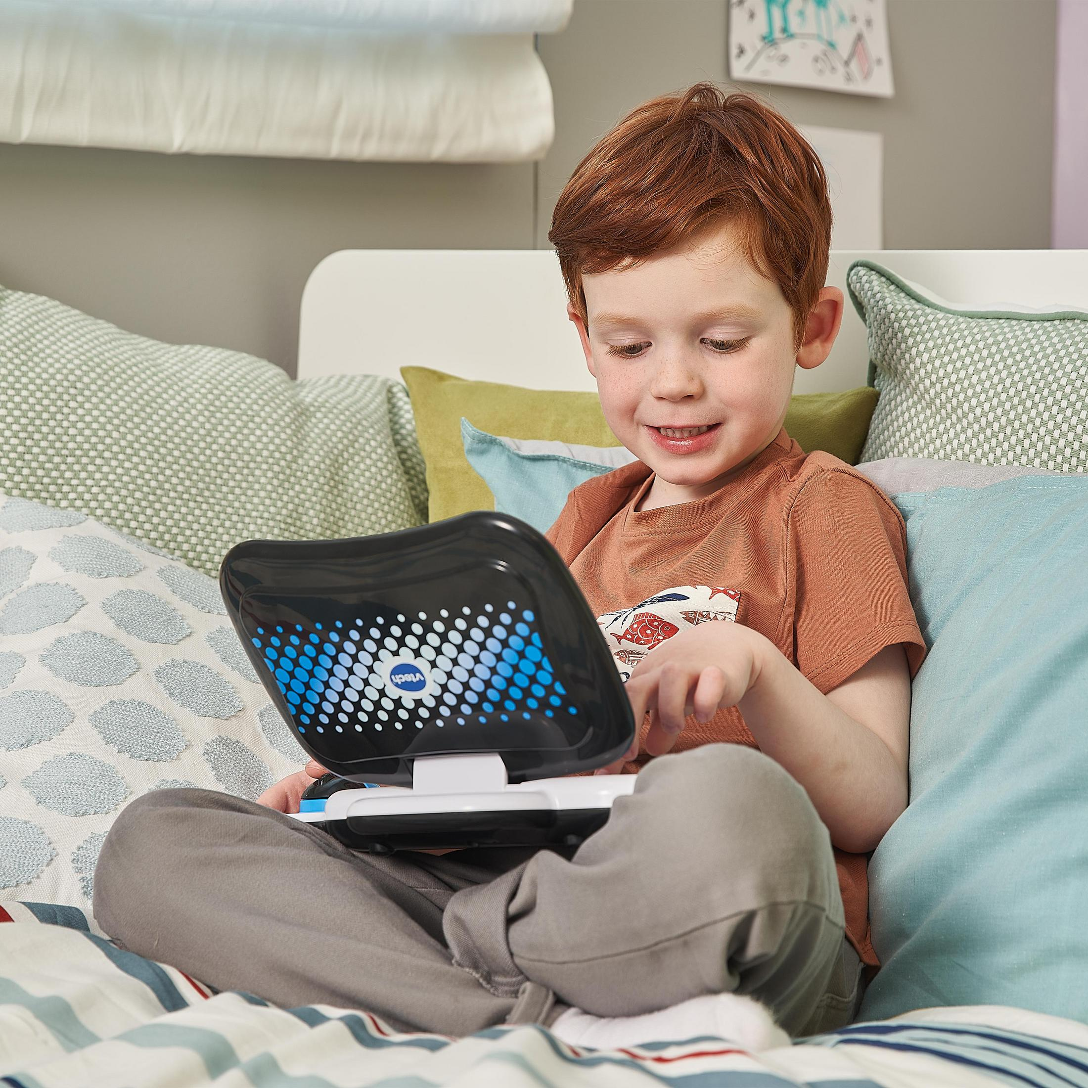 Ragazzo con i capelli rossi usa un laptop giocattolo seduto su un letto con cuscini.