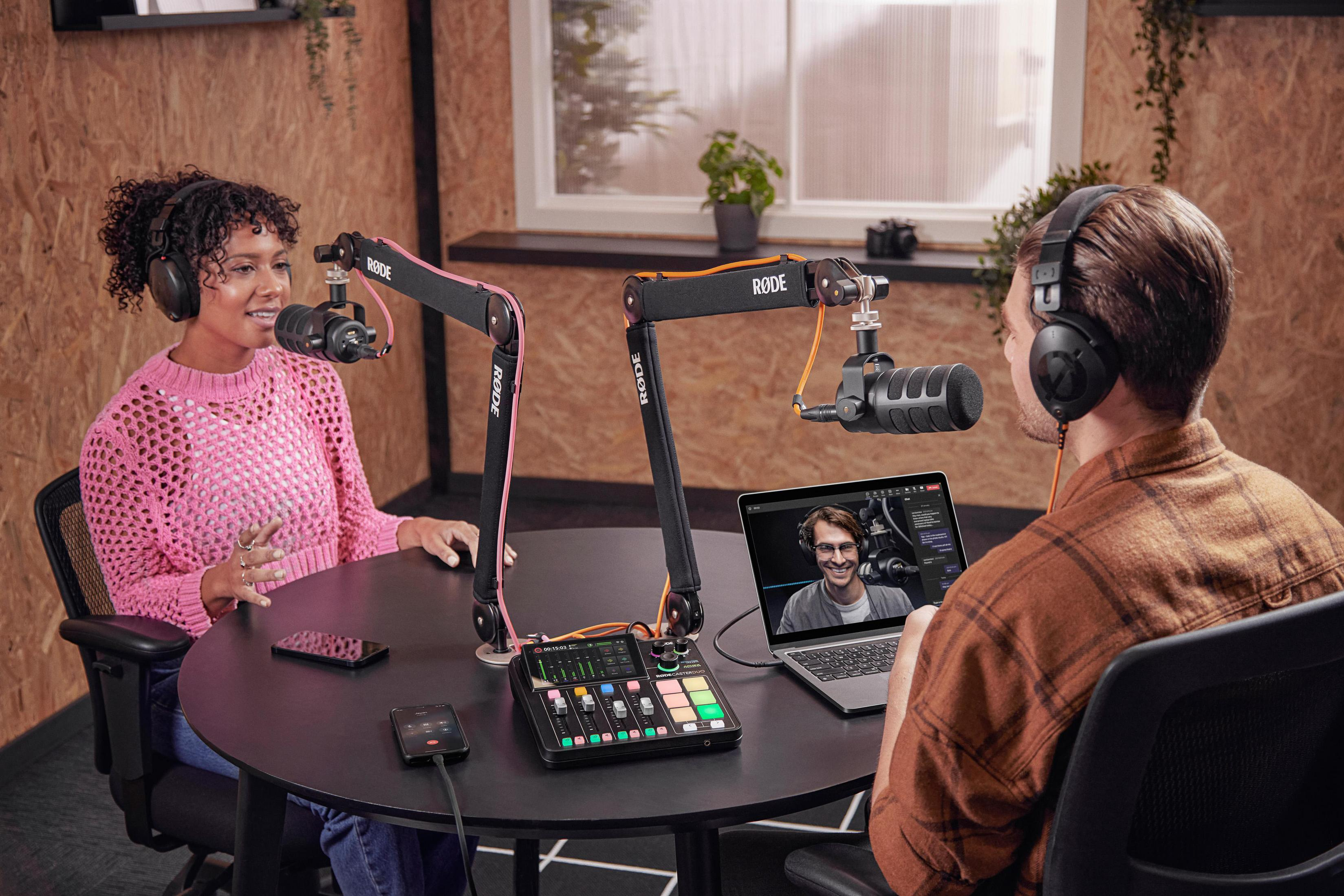 Due persone che fanno podcasting. Microfoni, mixer, laptop. Tavolo rotondo, sfondo con pannelli di legno.