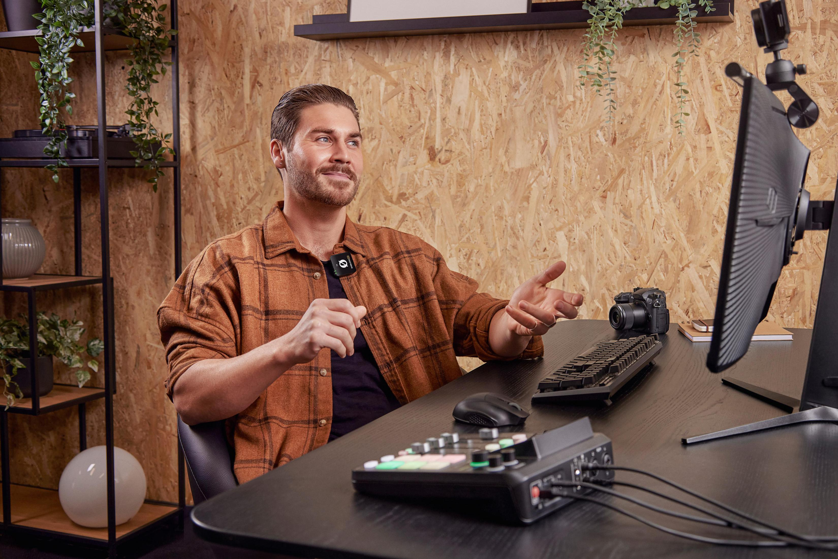 Un uomo con un microfono attaccato alla camicia seduto a una scrivania con un computer e un mixer audio.
