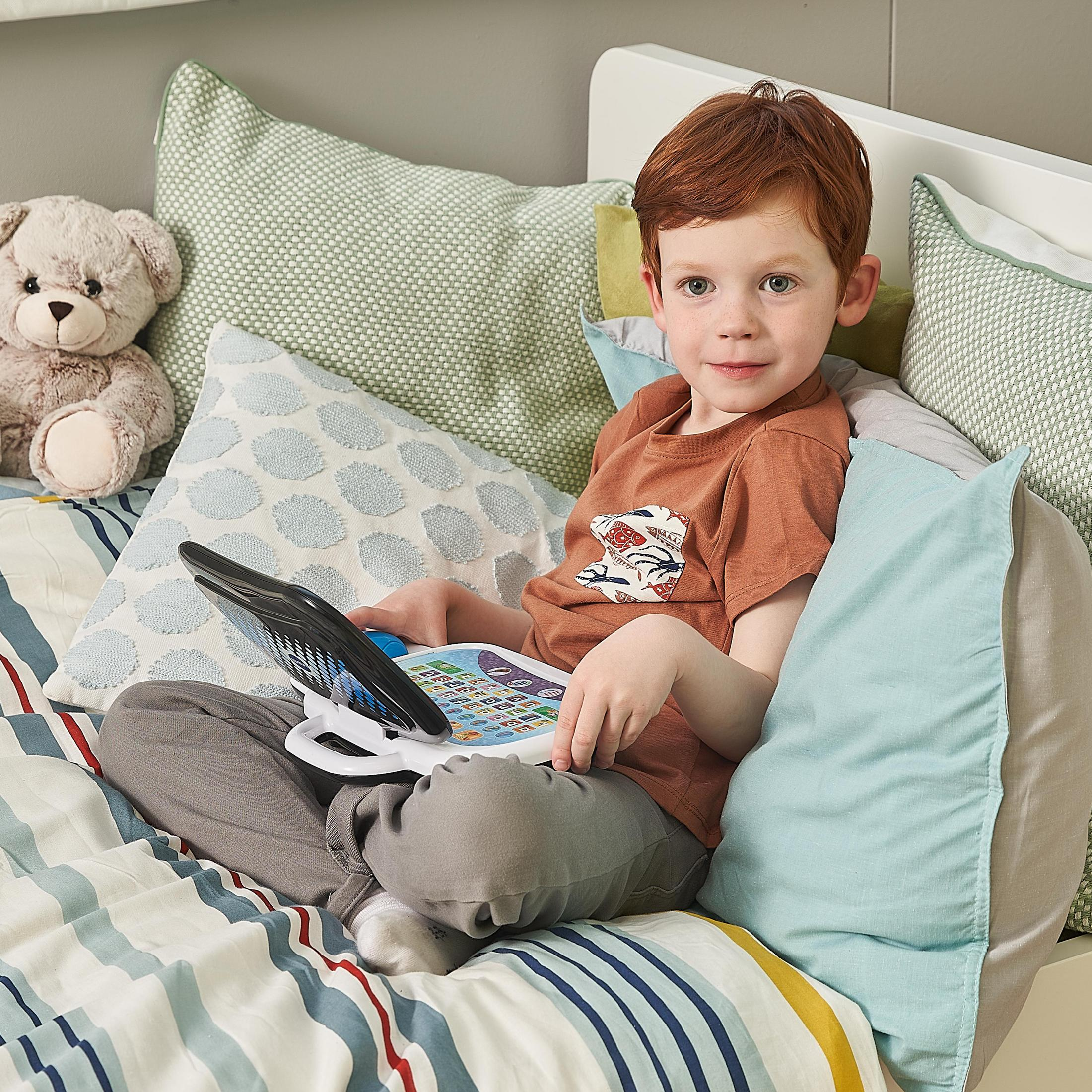 Un bambino tiene un laptop giocattolo sul letto. Sorride. C'è un orsacchiotto.