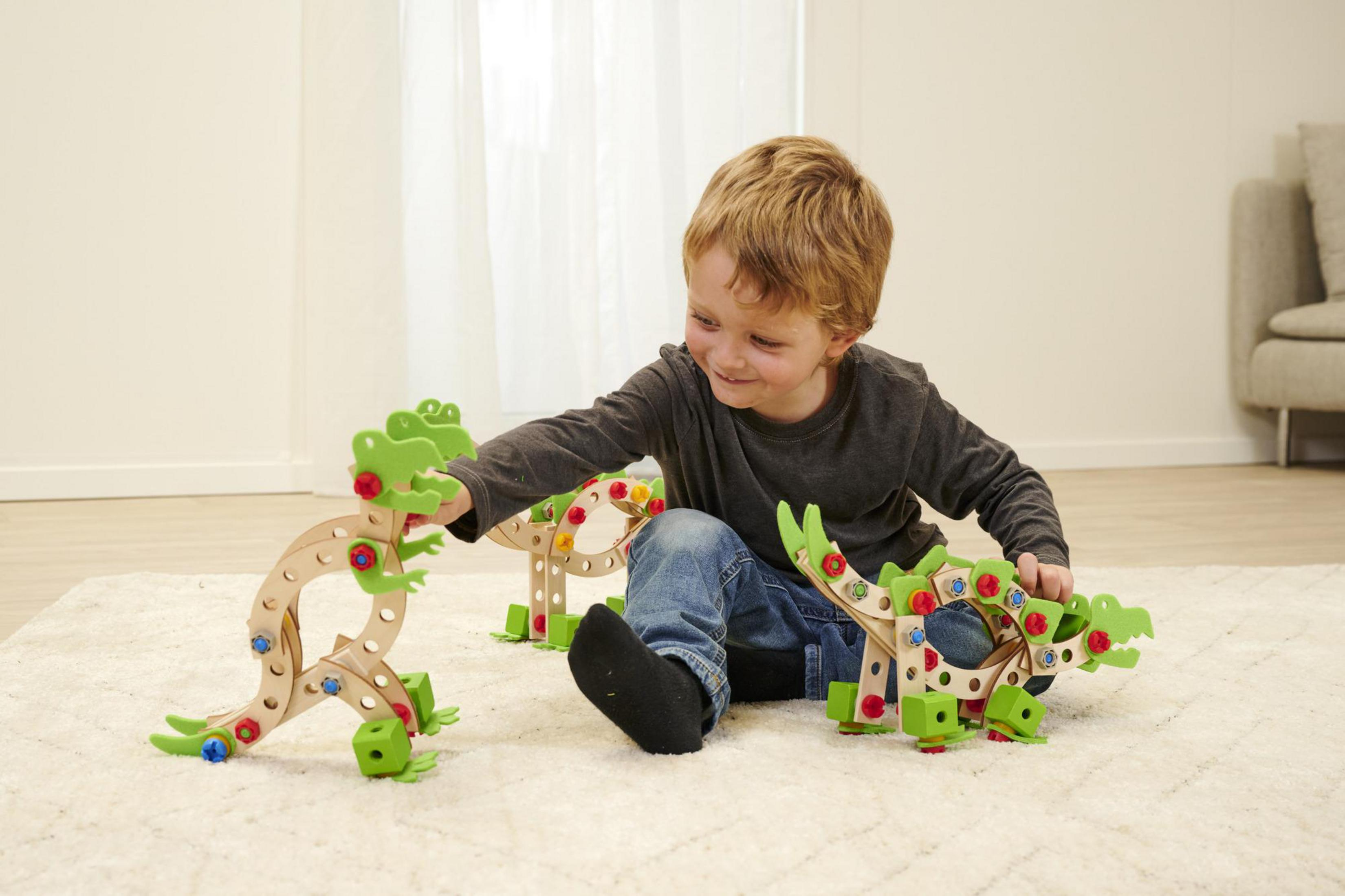 Ein lächelnder Junge spielt mit Holz-Dinosaurier-Spielzeugen auf einem Teppich. Zwei Dinosaurier werden gebaut.