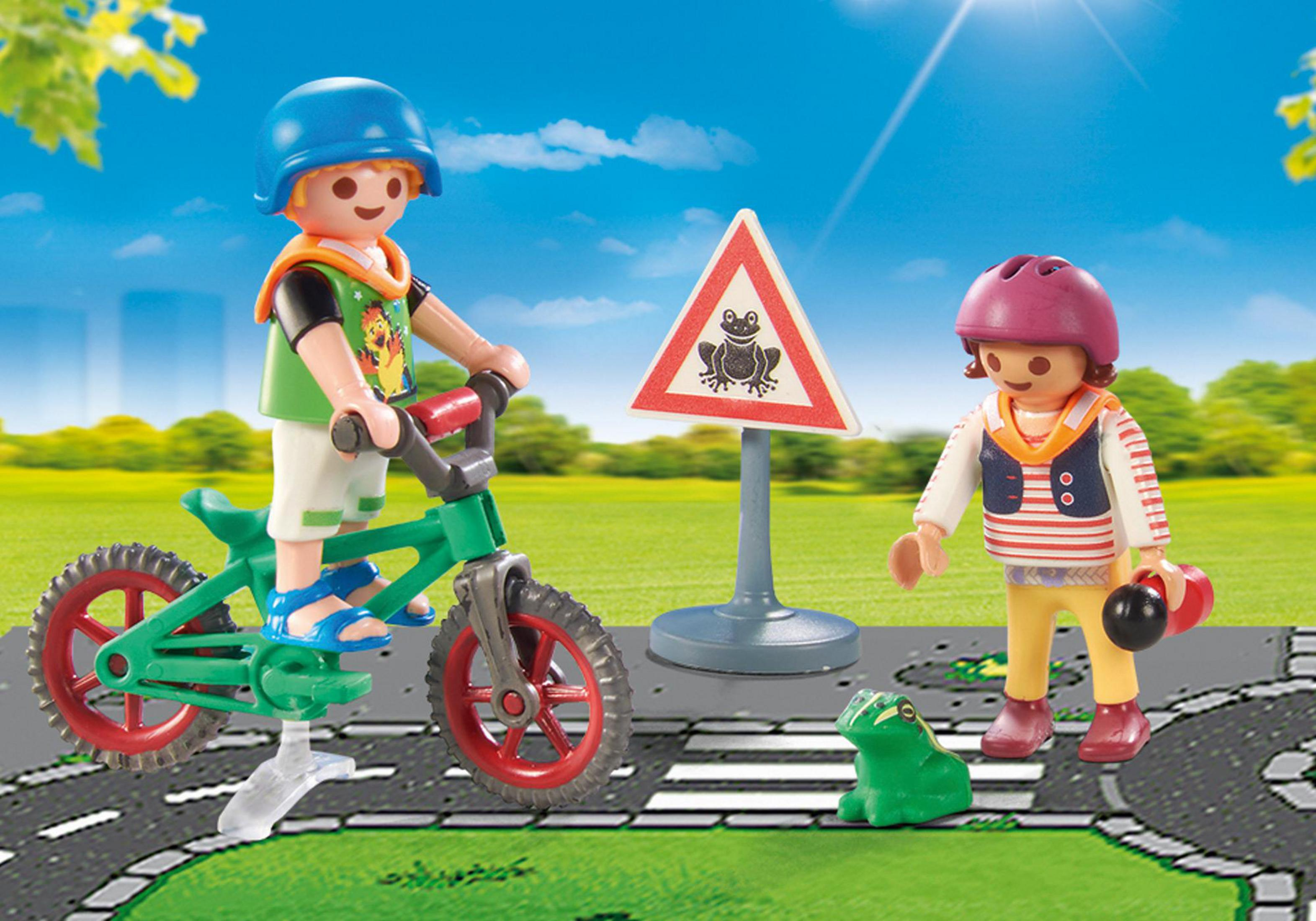 Zwei Playmobil-Figuren mit Fahrrad und Frosch-Zeichen auf einer Straße. Grünes Gras und blauer Himmel.