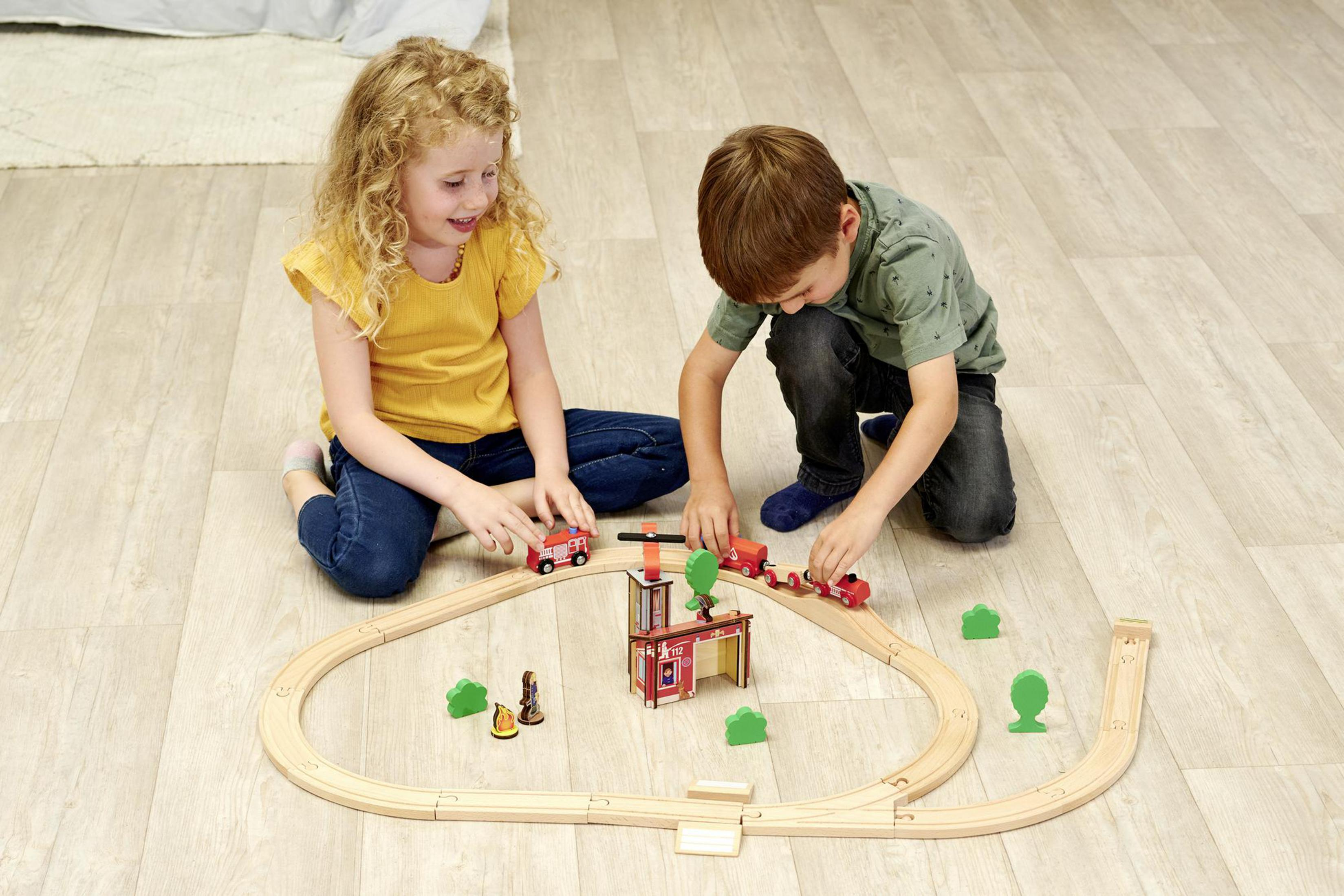 Dos niños jugando con un tren de juguete. Vías de madera, trenes rojos y verdes.