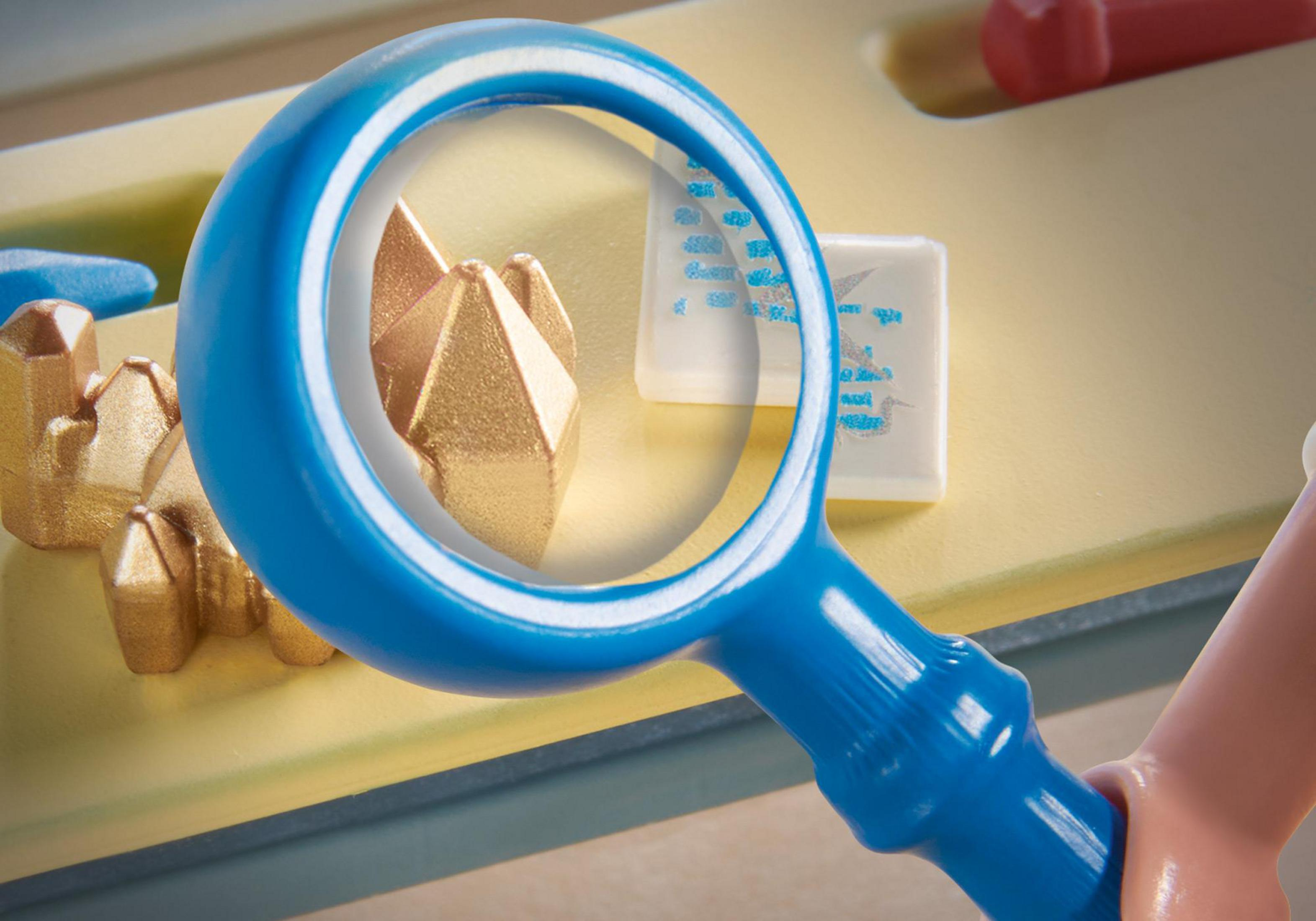 Lupa skupia się na złotych kryształach na żółtej powierzchni, z innymi obiektami rozmytymi w tle.