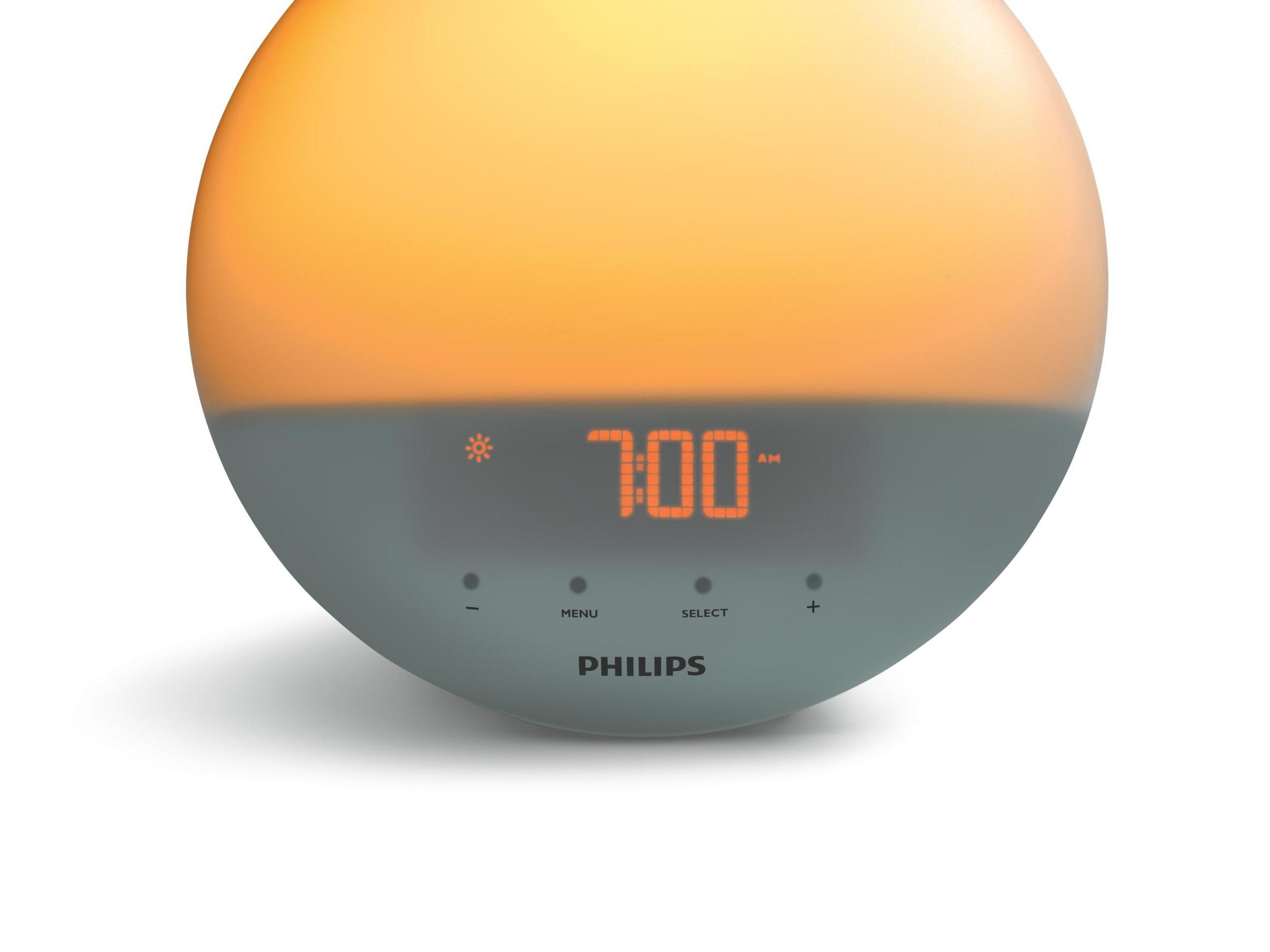 Une lampe de réveil Philips ronde et grise affiche 7h00 du matin sur un écran. Les boutons en dessous indiquent MENU, SELECT et +
