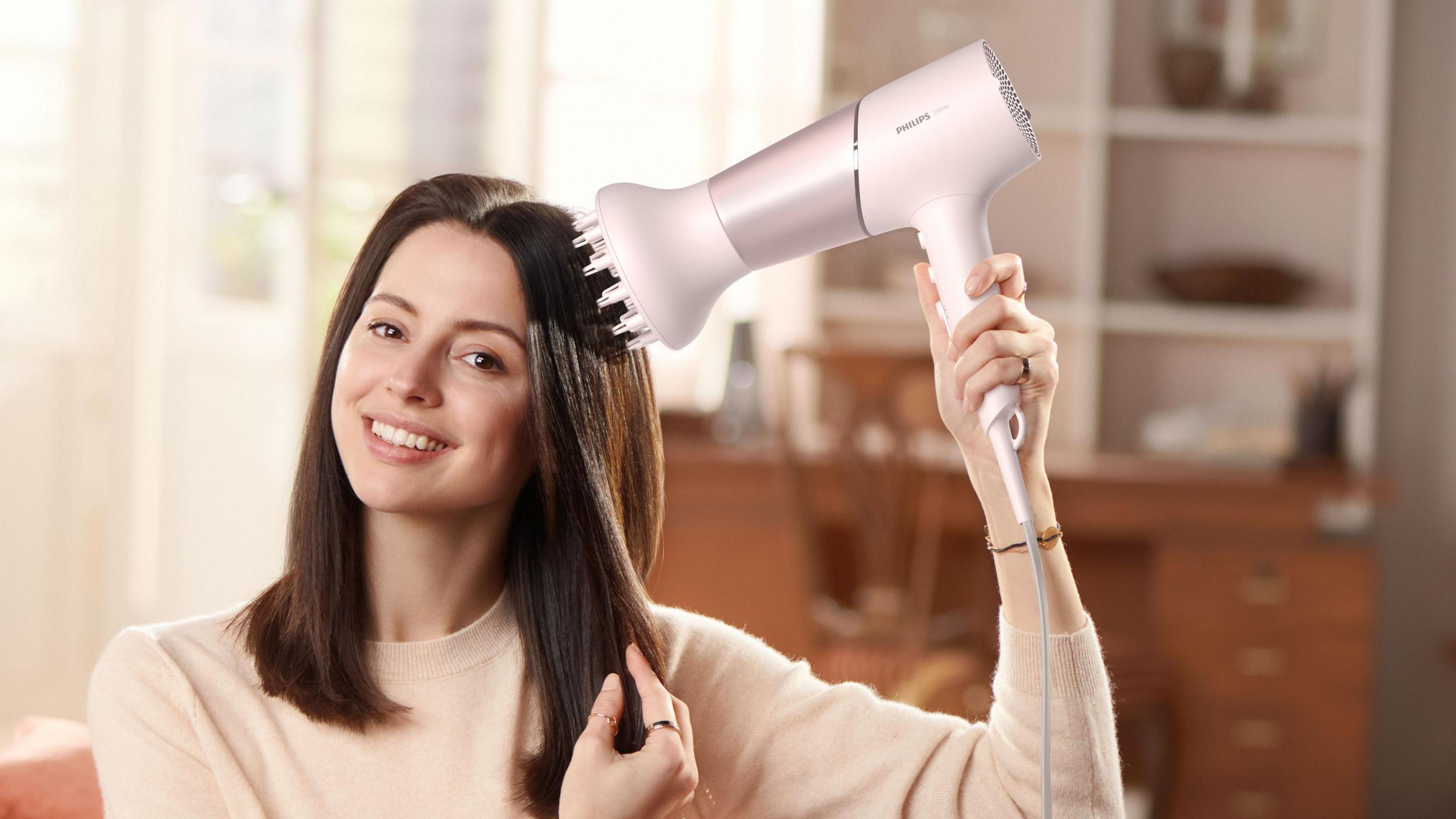 Mujer usando un secador de pelo rosa en su cabello oscuro, sonriendo en interiores.