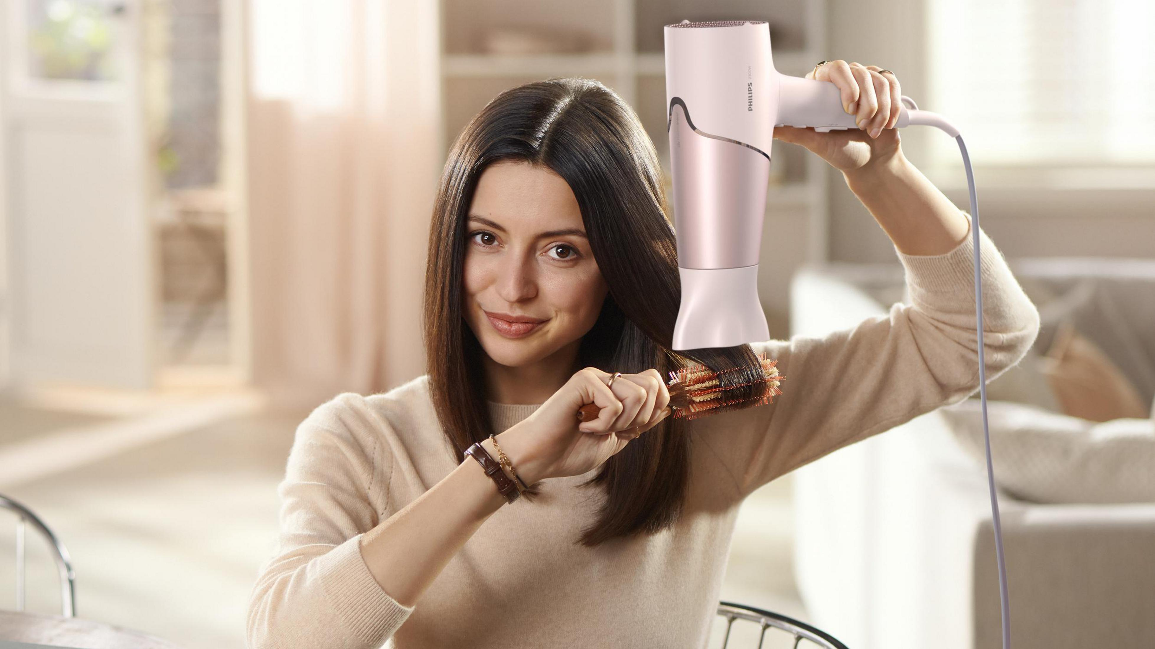 Mujer usando secador y cepillo. Sonríe peinándose. El secador es rosa.