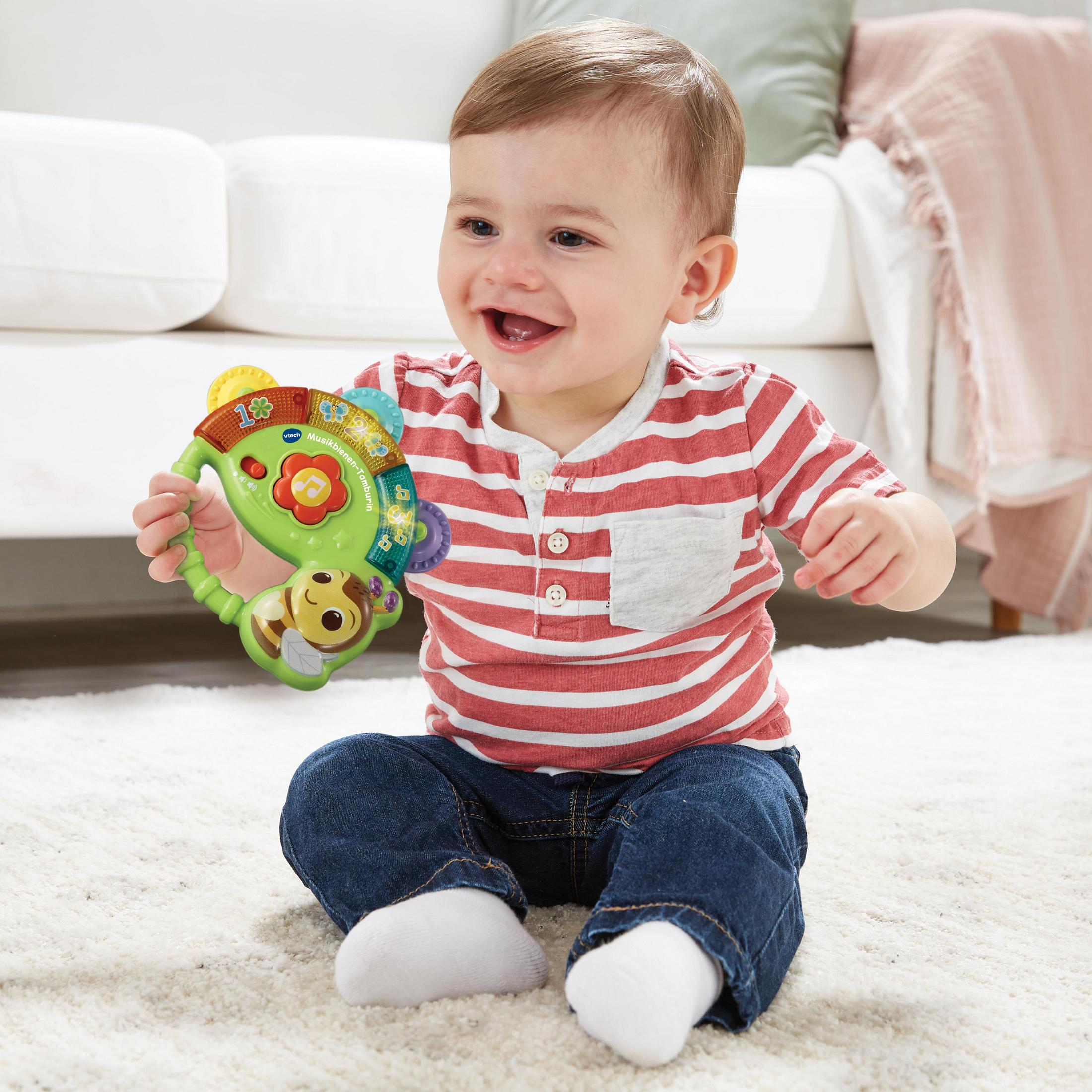Ein Baby lacht und hält ein grünes Spielzeug mit einem Affen und einer Blume. Das Baby sitzt auf einem Teppich.