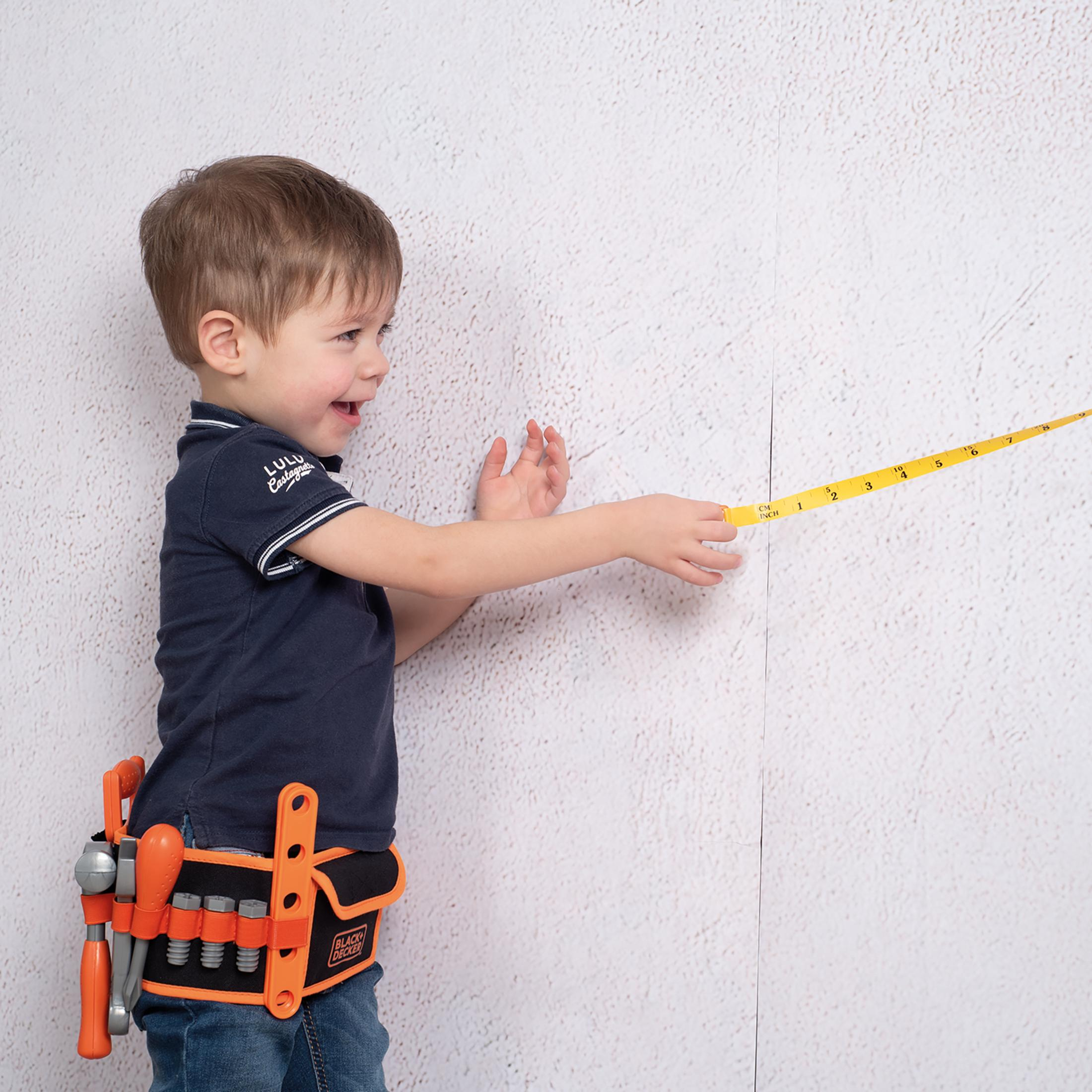 Ein kleiner Junge mit Werkzeuggürtel misst mit einem gelben Maßband eine Wand. Er lächelt, die Wand ist weiß.