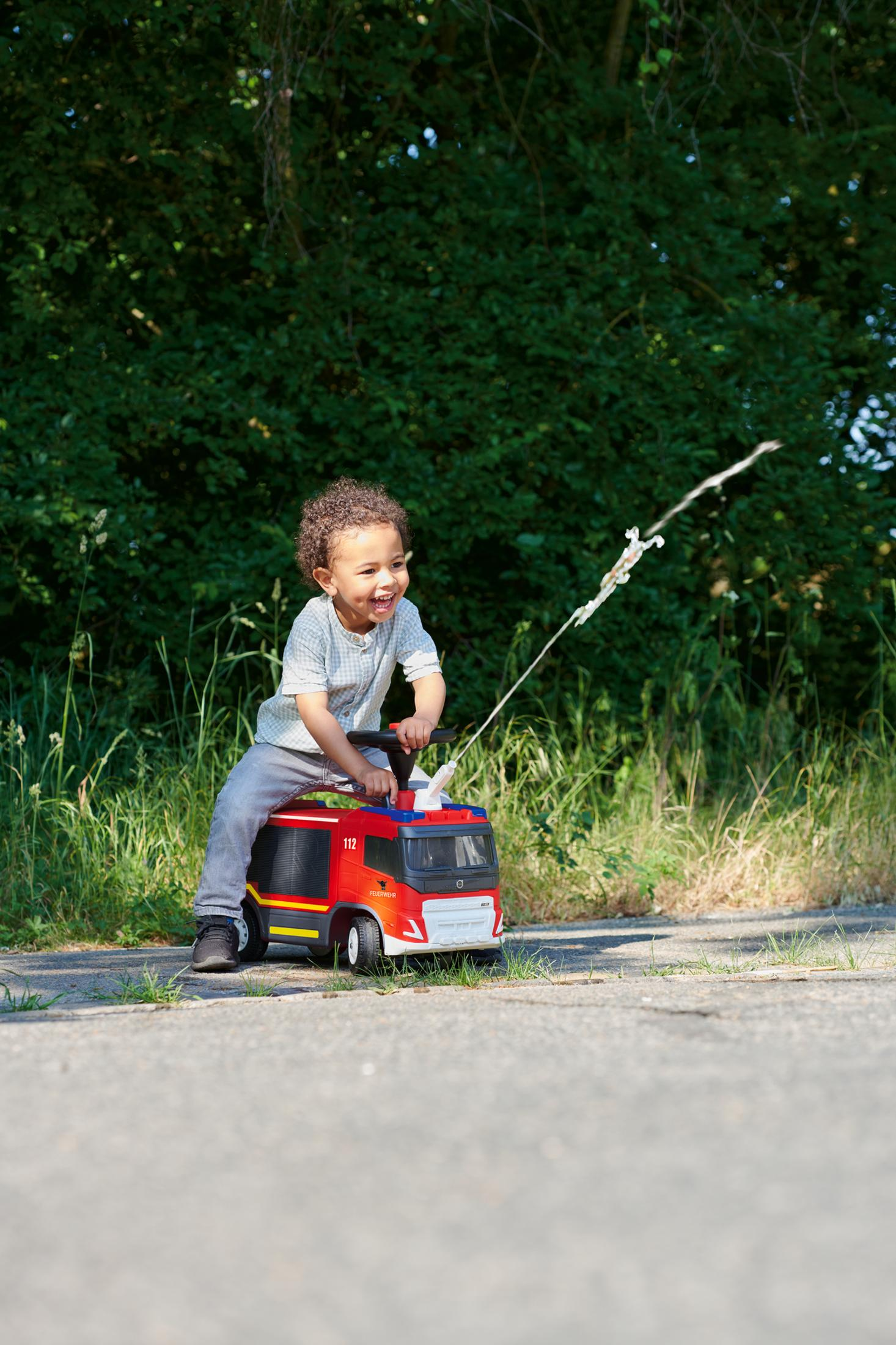 Junge auf Spielzeug-Feuerwehr spritzt Wasser. Grüne Bäume und eine Straße im Hintergrund.