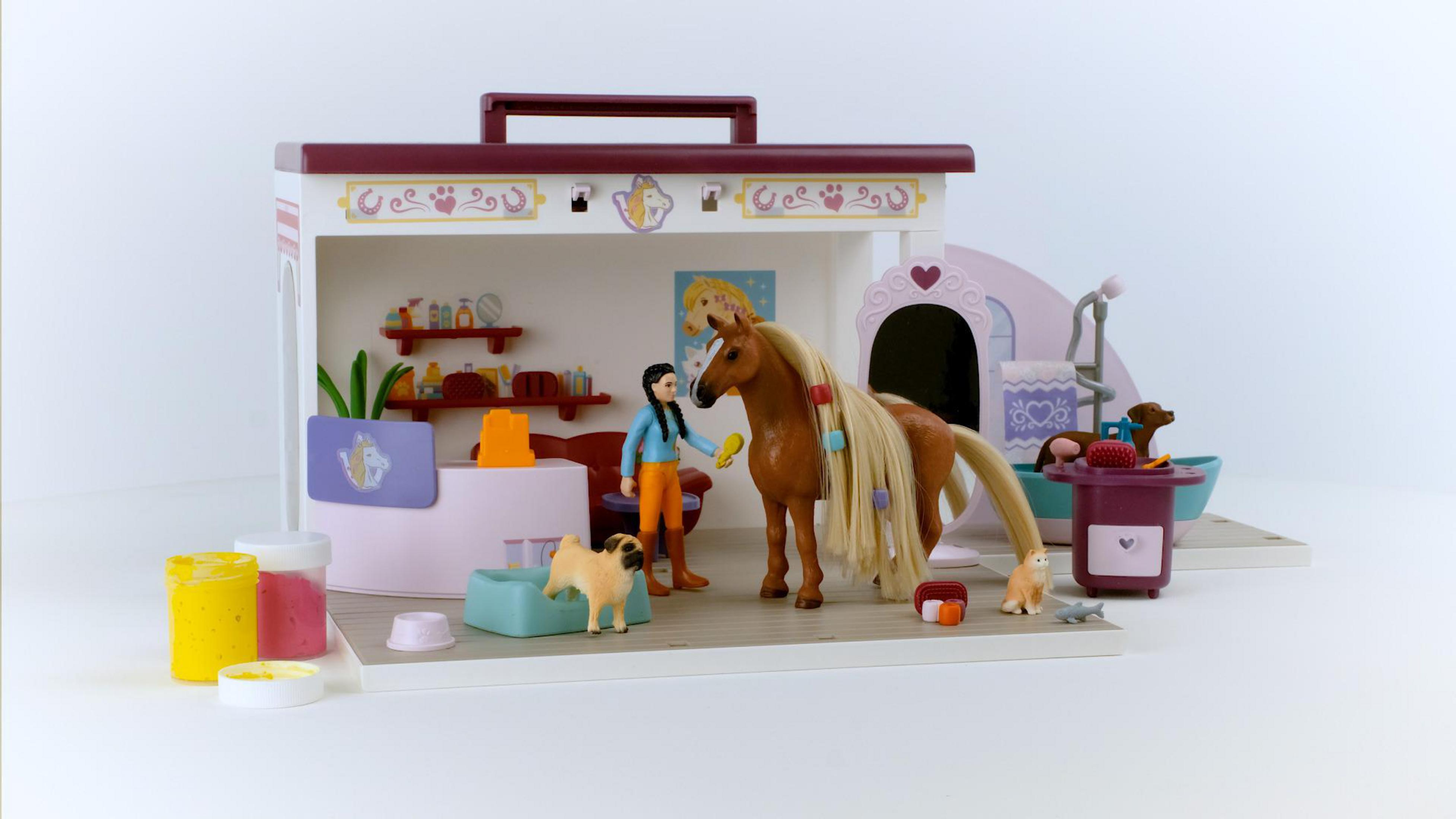 Spielzeug-Tierpflege-Set mit einer Person, die ein Pferd, einen Hund und Zubehör pflegt. Der Hintergrund ist weiß.