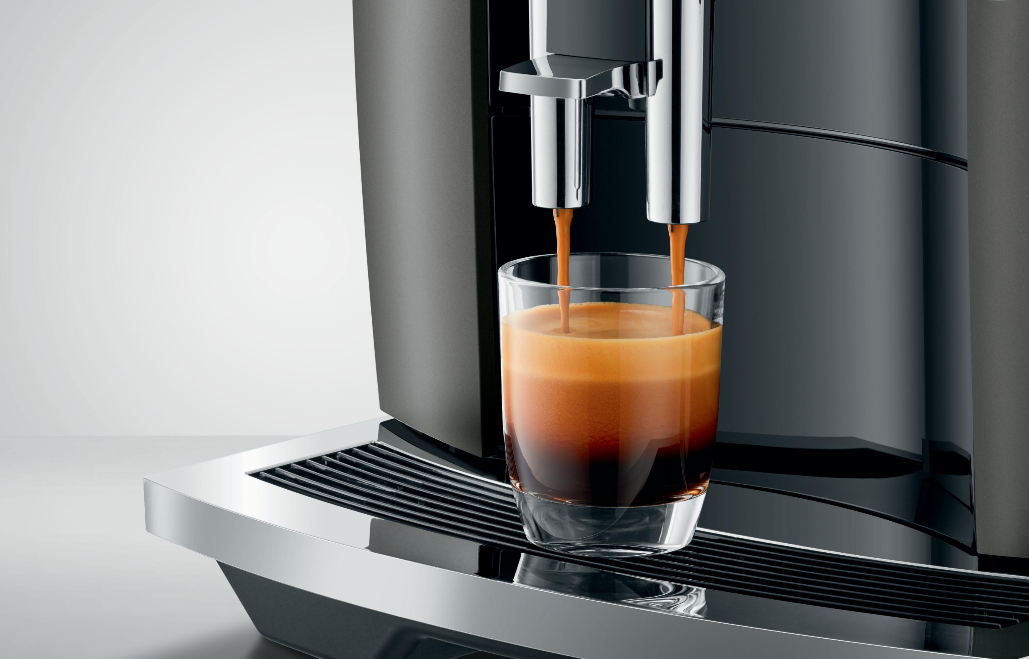 Espresso, der aus einer schwarzen Kaffeemaschine in ein klares Glas gegossen wird. Das Glas steht auf einem Metalltablett.