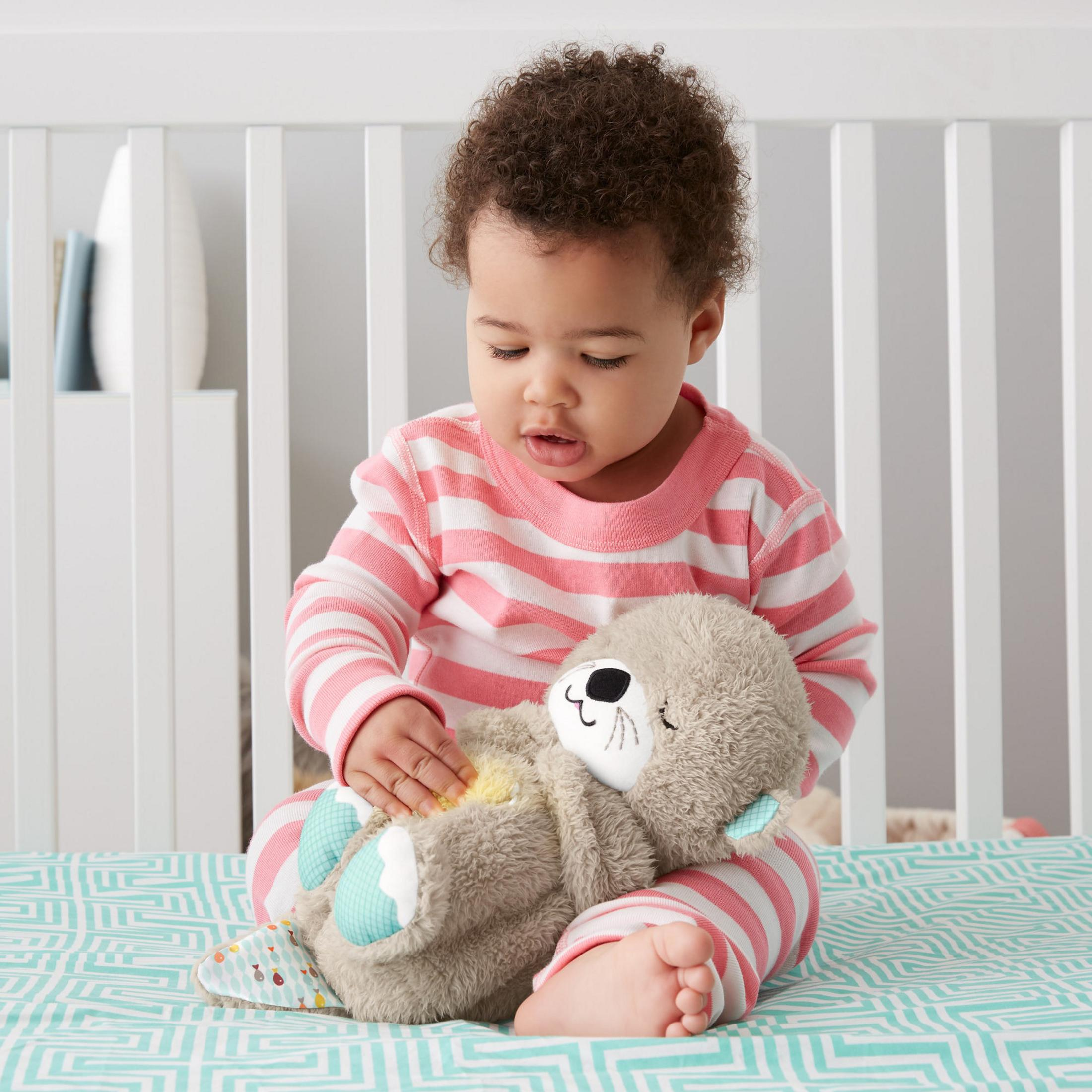 Baby spielt mit einem Stoffotter auf einem Bett. Rosa-weiß gestreifter Schlafanzug und ein Kinderbett.