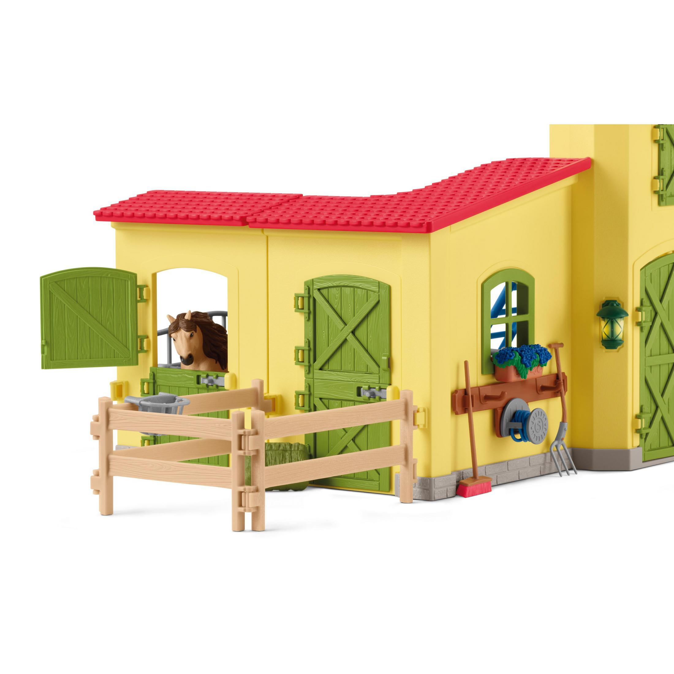 Gelber Spielzeugstall mit grünen Türen und Dach. Ein Pferd ist in einem Stall. Zaun, Trog und Werkzeuge sichtbar.
