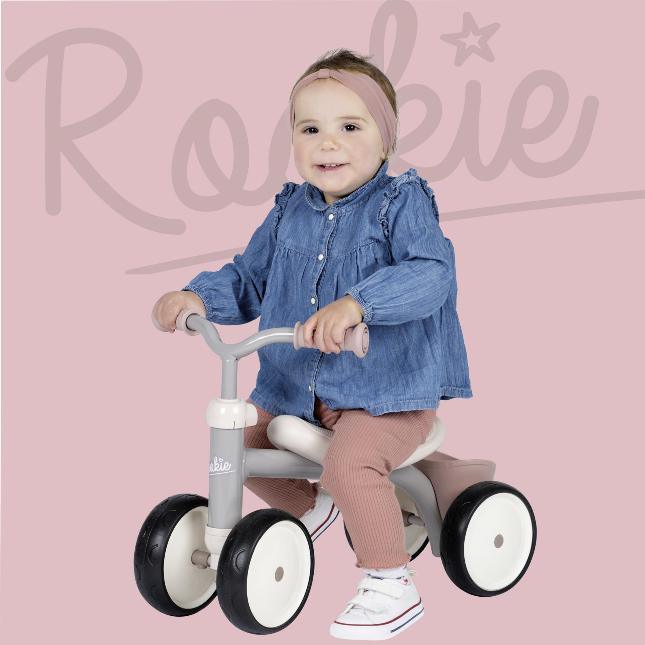 Ein Baby auf einem Dreirad lächelt, trägt ein Stirnband. Rosa Hintergrund mit 'Rookie'-Text.