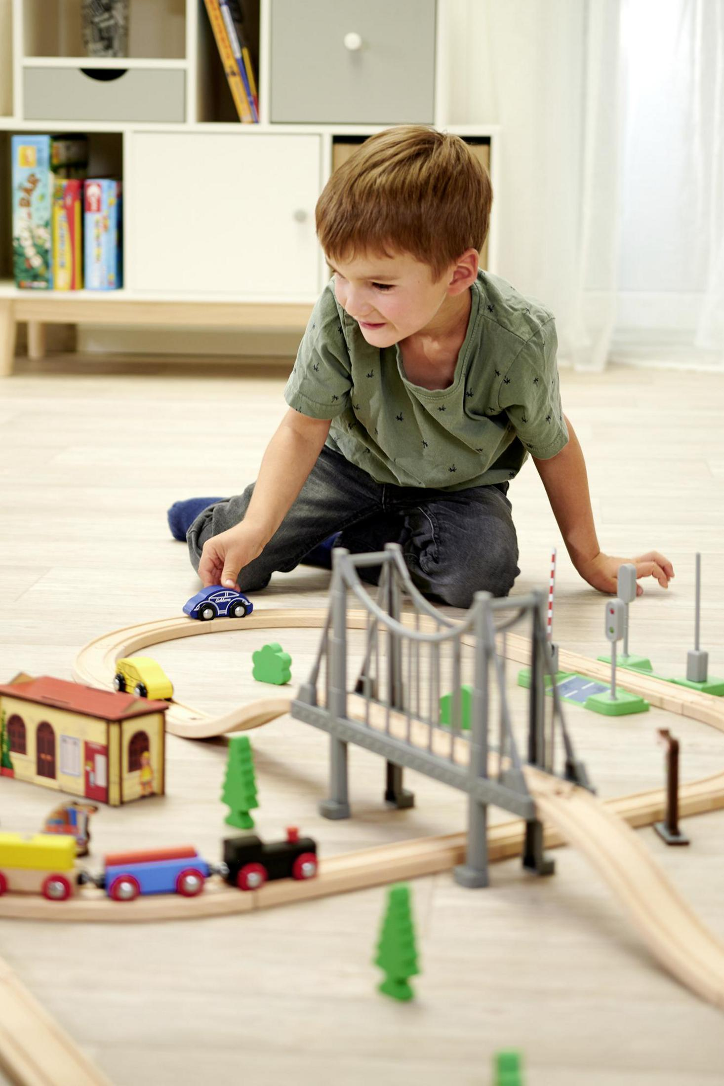 Ein Junge spielt mit einer Spielzeugeisenbahn auf dem Boden. Eine Spielzeugeisenbahn und eine Brücke sind sichtbar.