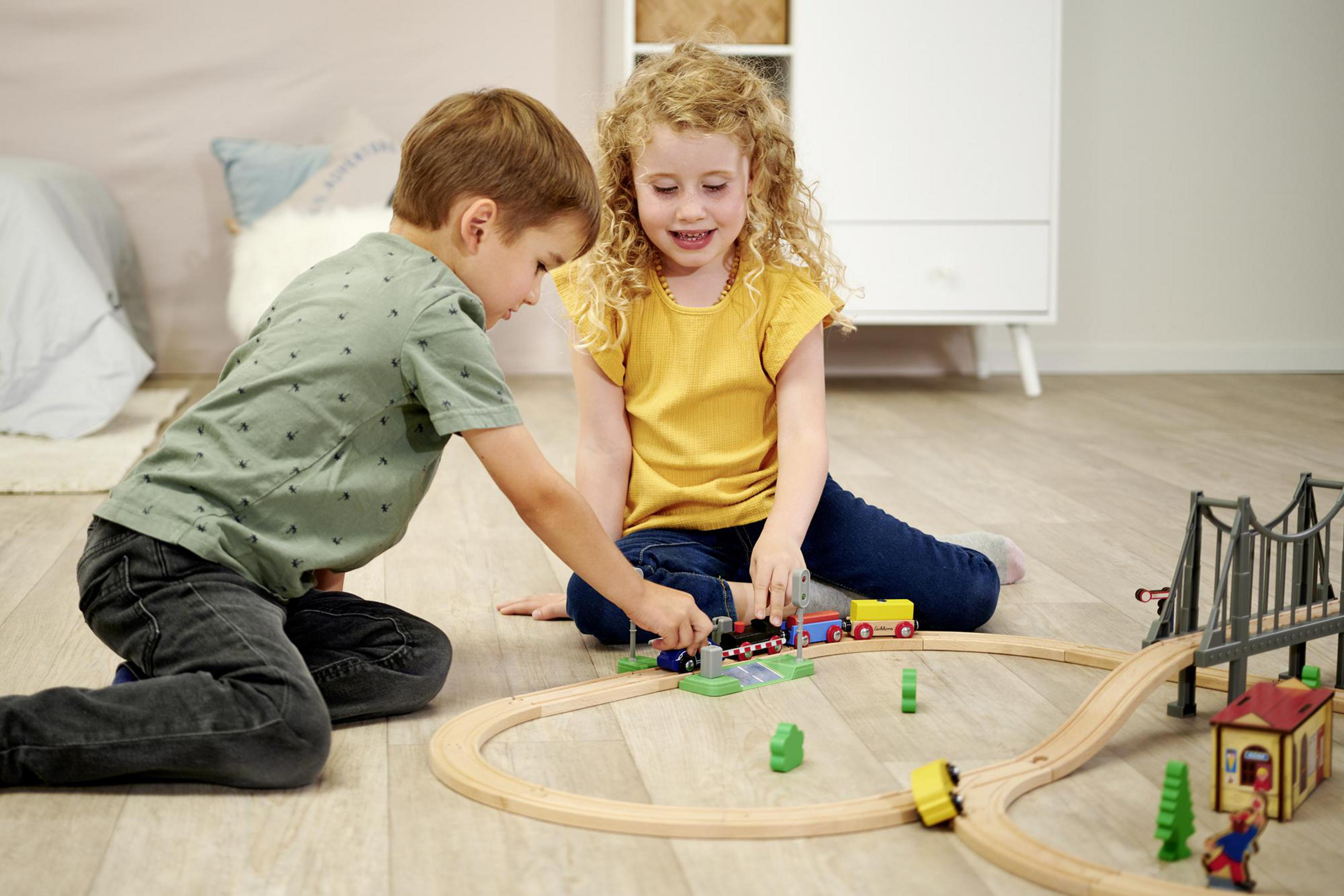 Zwei Kinder spielen mit einem Holzeisenbahn-Set auf einem Holzboden. Eine Brücke ist vorhanden.