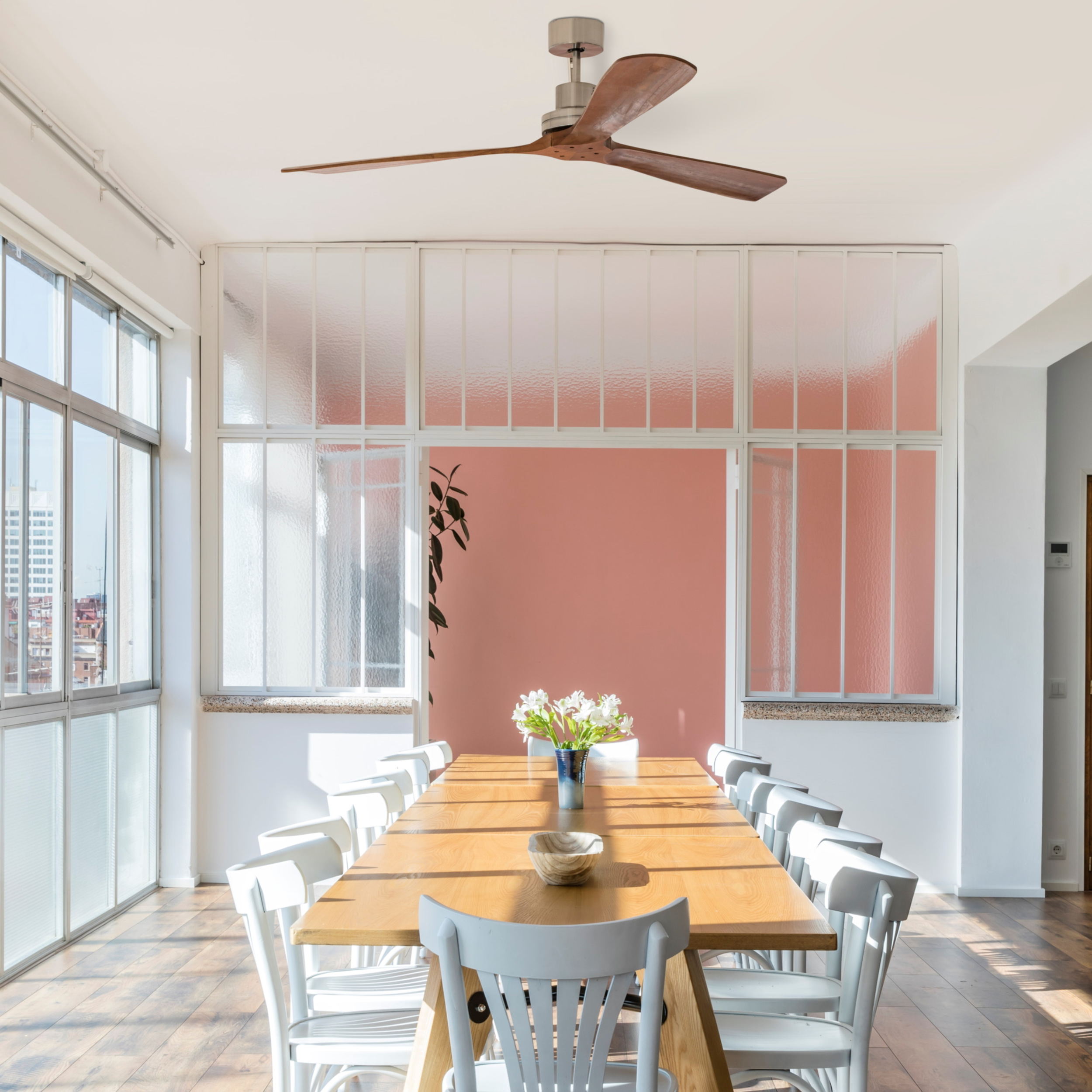 Esszimmer mit langem Holztisch, weißen Stühlen, großen Fenstern und Deckenventilator. Rosa Wand im Hintergrund.