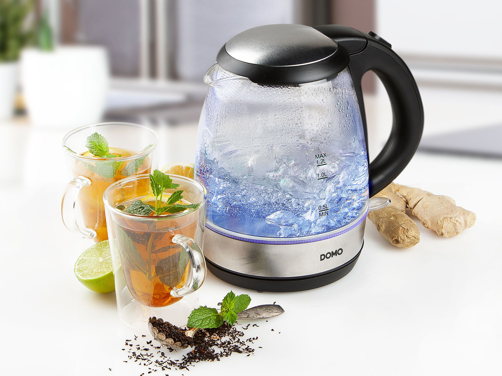 Szklany czajnik elektryczny z wrzącą wodą, dwiema szklankami herbaty i plasterkami imbiru.