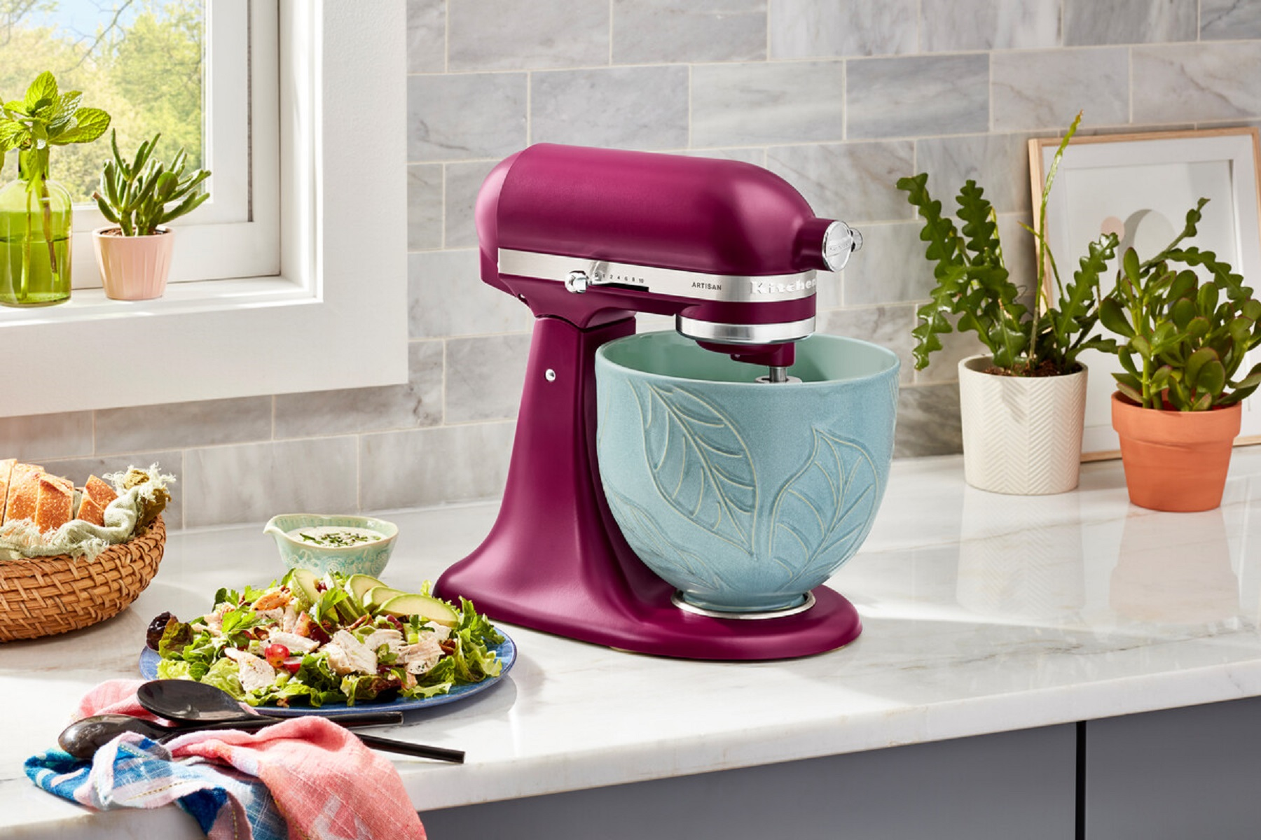 Ein magentafarbener KitchenAid-Mixer mit hellblauer Schüssel, umgeben von Essen und Pflanzen.