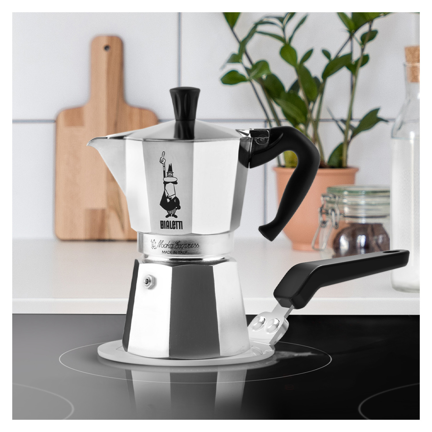 Una cafetera Moka Bialetti plateada está sobre una estufa negra. Una tabla de cortar de madera y plantas cerca.