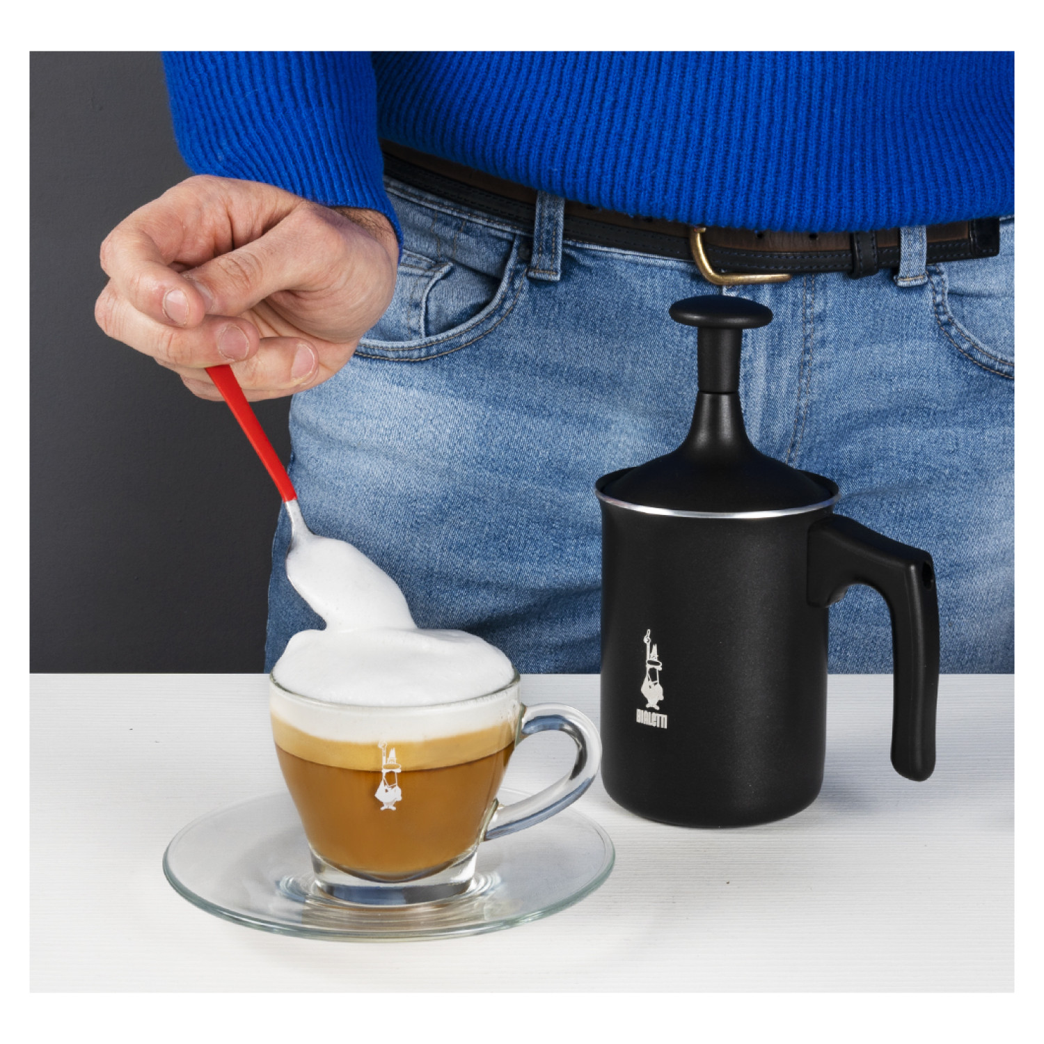 Osoba robiąca kawę. Filiżanka kawy z pianką obok czarnego spieniacza.