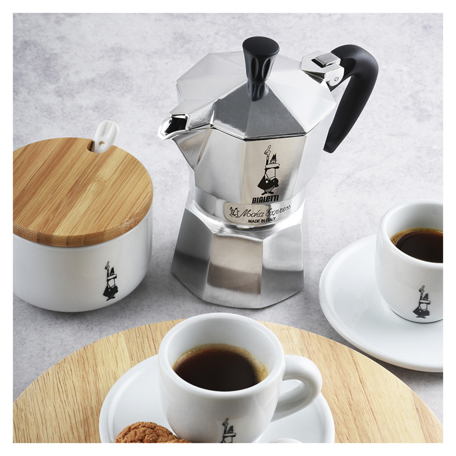 Srebrny ekspres do espresso, cukiernica i dwie filiżanki kawy na drewnianej tacy.