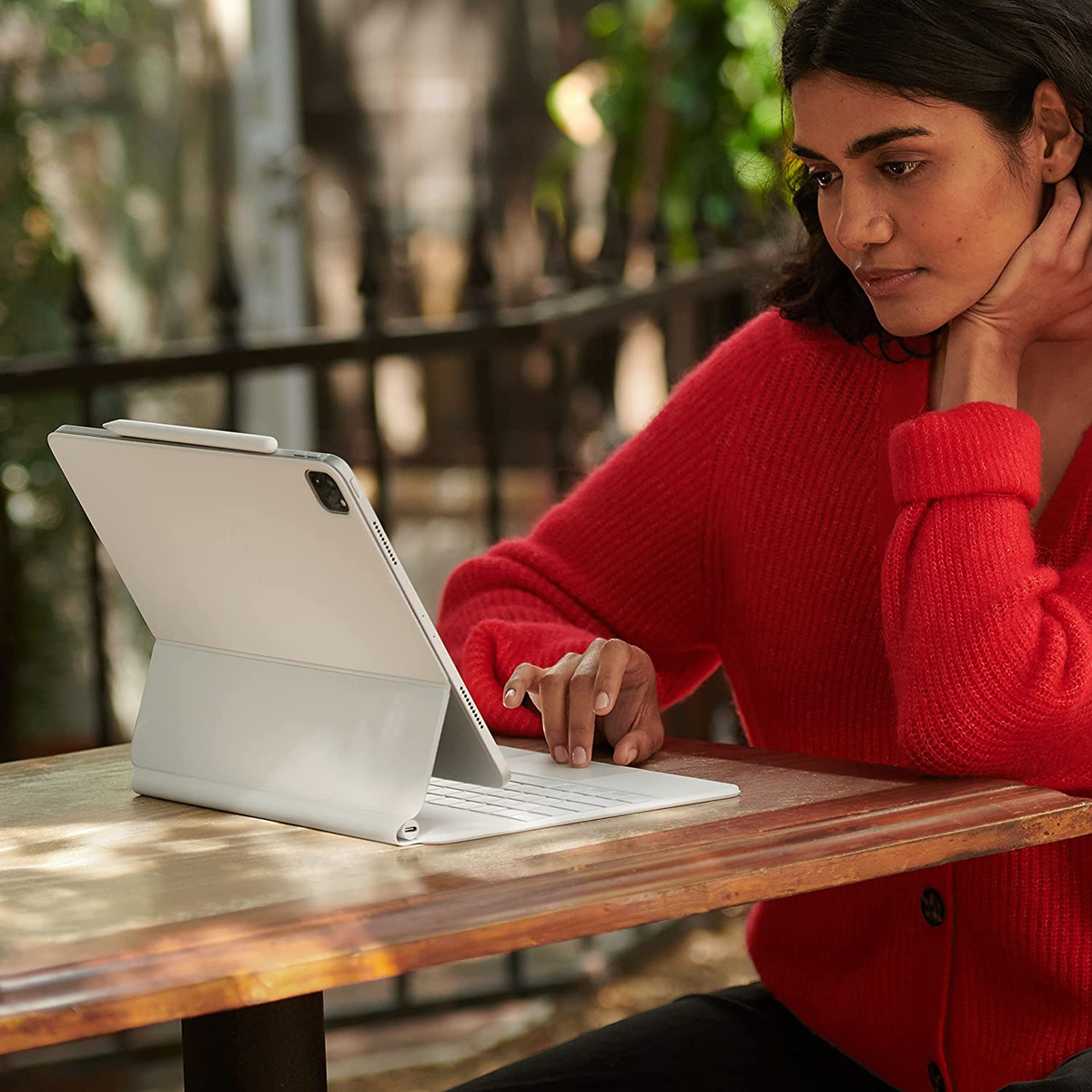 Eine Frau sitzt an einem Tisch und benutzt ein Tablet mit Tastatur. Sie trägt einen roten Pullover. Hintergrund draußen.