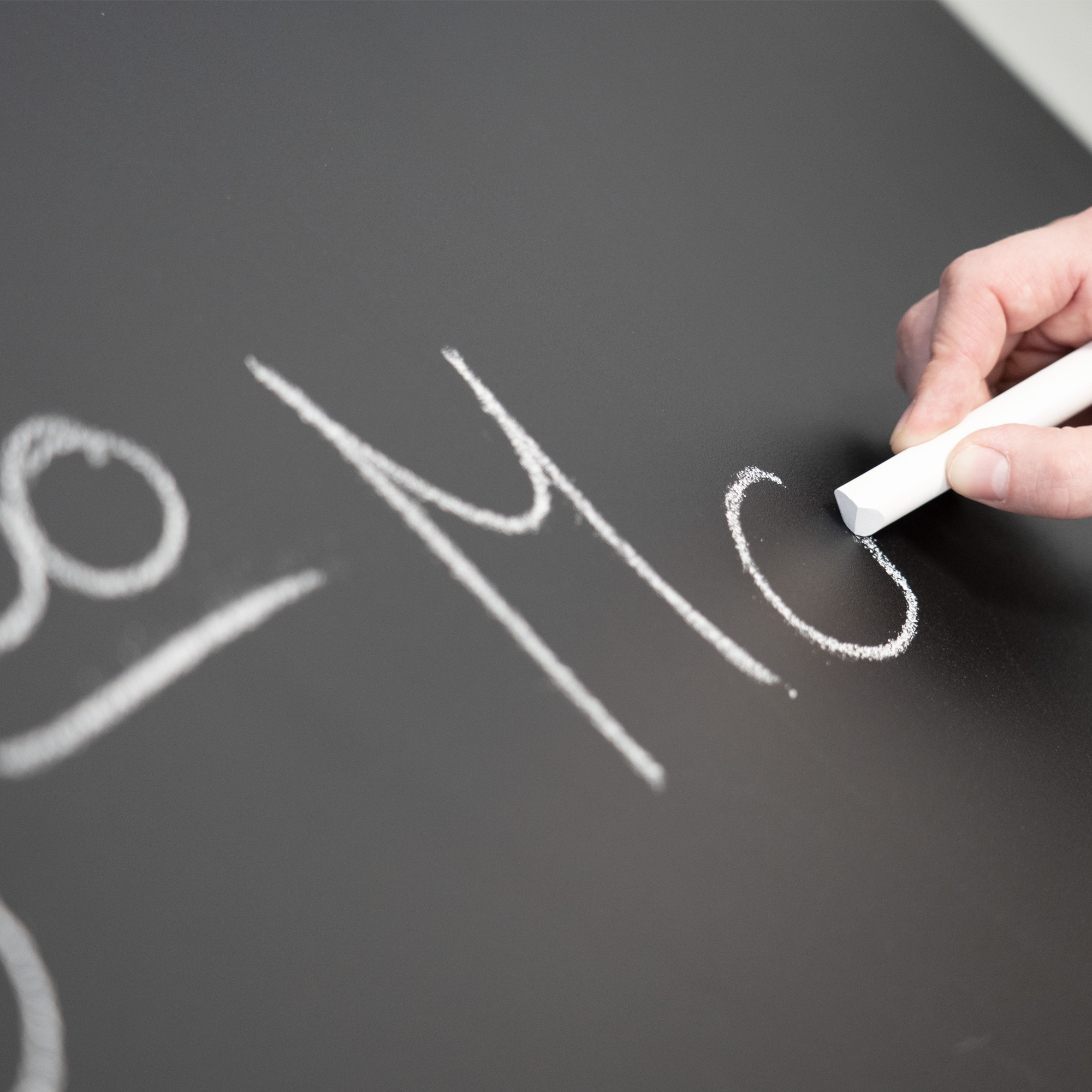 Hand schreibt mit Kreide den Buchstaben 'M' auf eine schwarze Oberfläche.