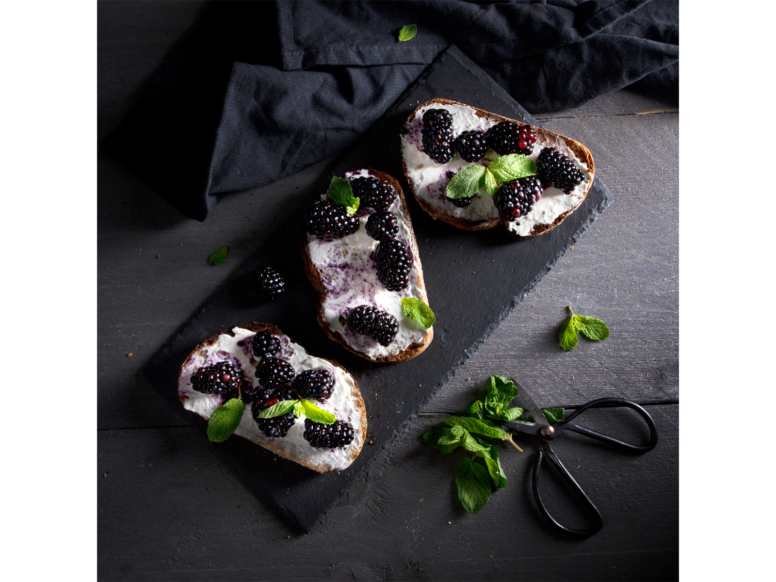 Trzy kromki chleba z kremowym serem i jeżynami na czarnej łupku z miętą.