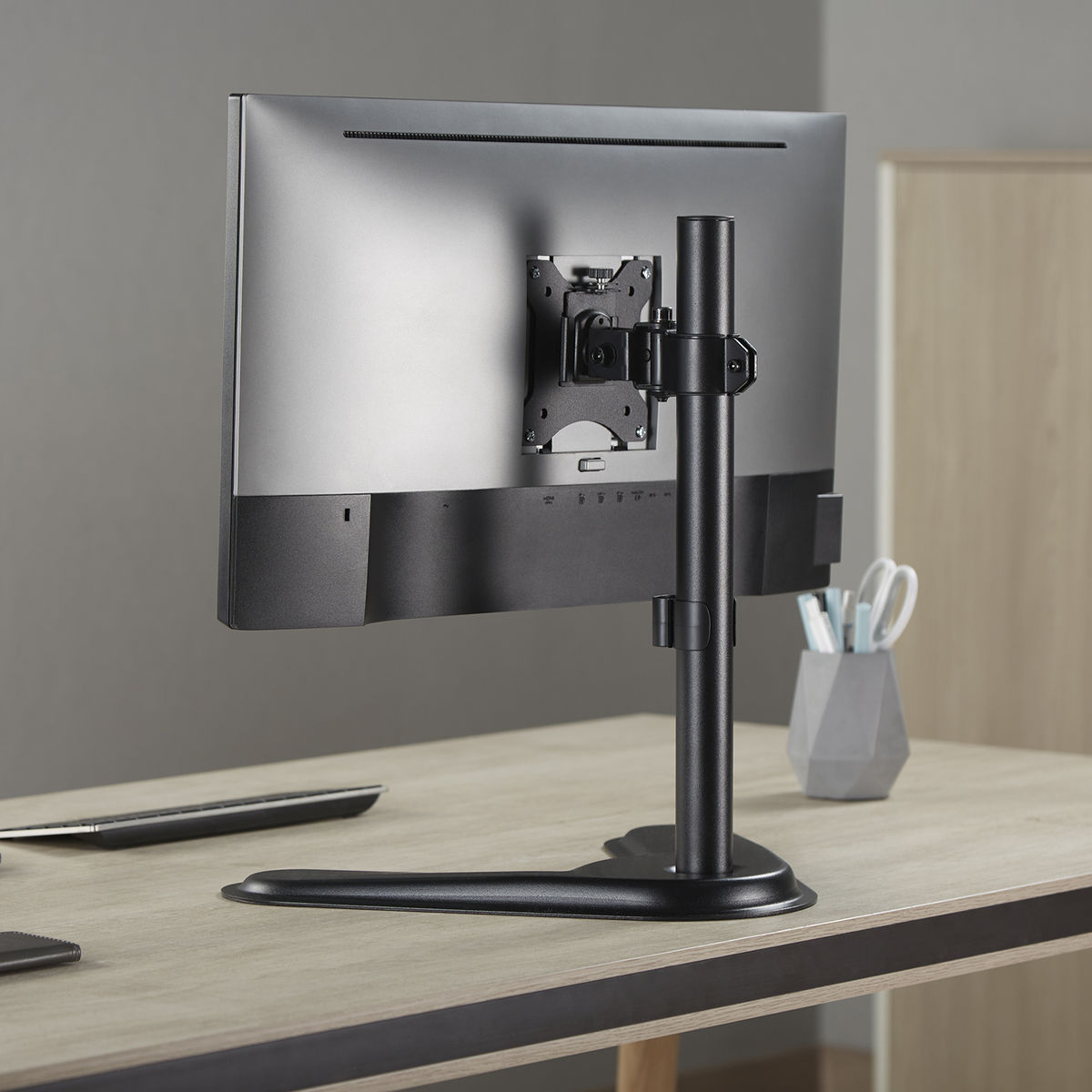 Een monitor op een standaard op een bureau. De achterkant van de monitor is zichtbaar, met toetsenbord en penhouder.
