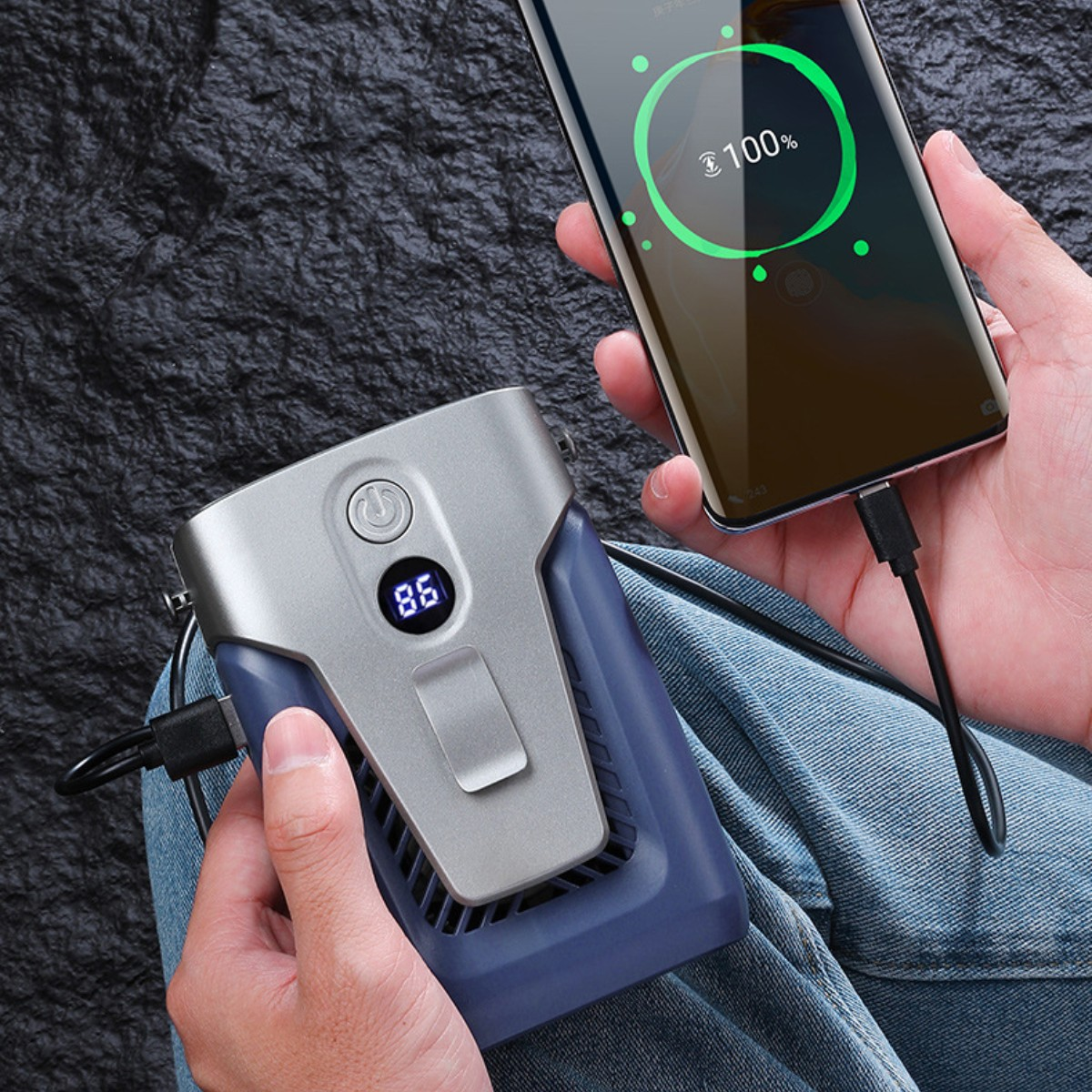 Eine Person hält einen tragbaren Ventilator und ein Smartphone, das geladen wird. Dunkler Hintergrund.