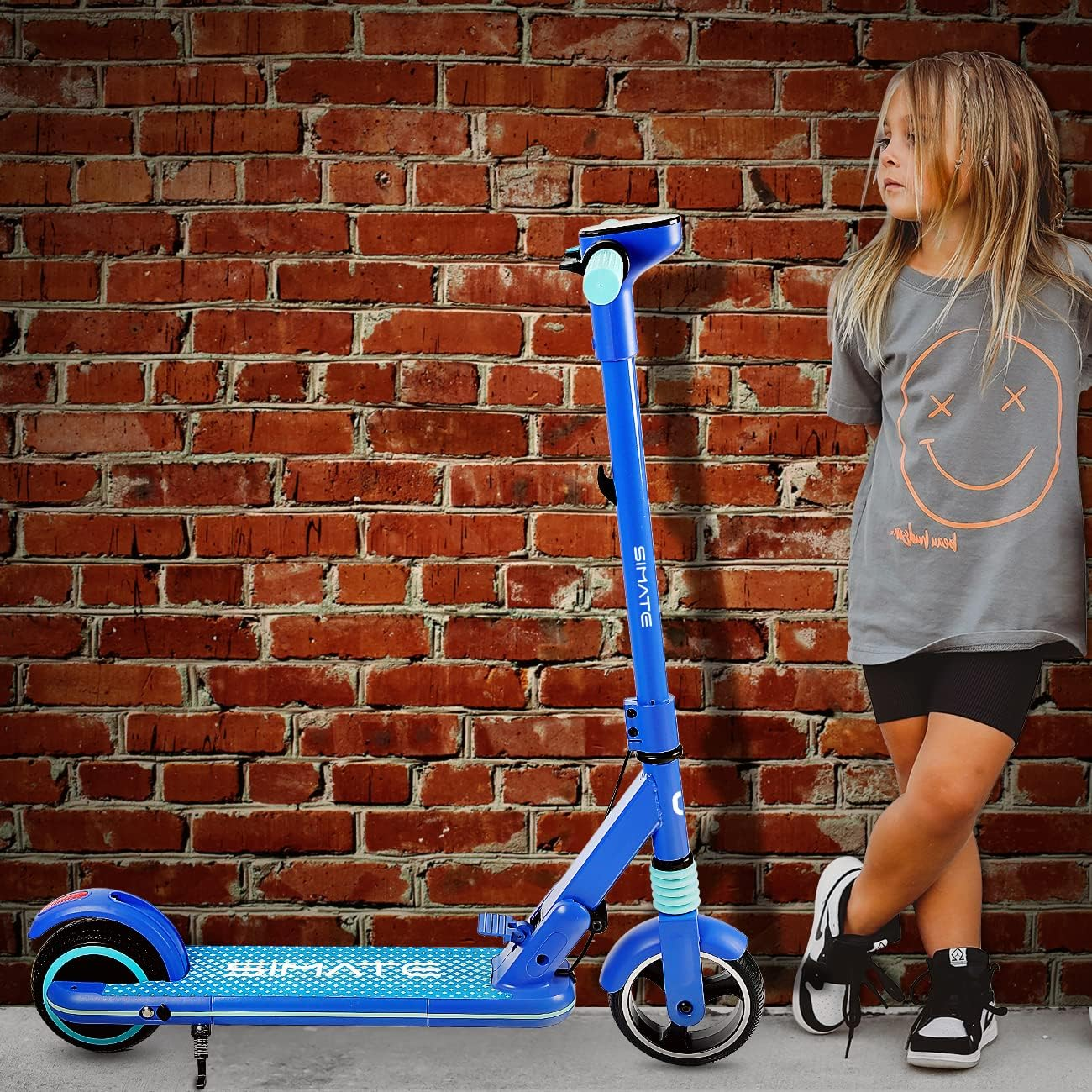 Junges Mädchen mit blauem Roller, lehnt an einer Backsteinmauer mit Smiley-Grafik.