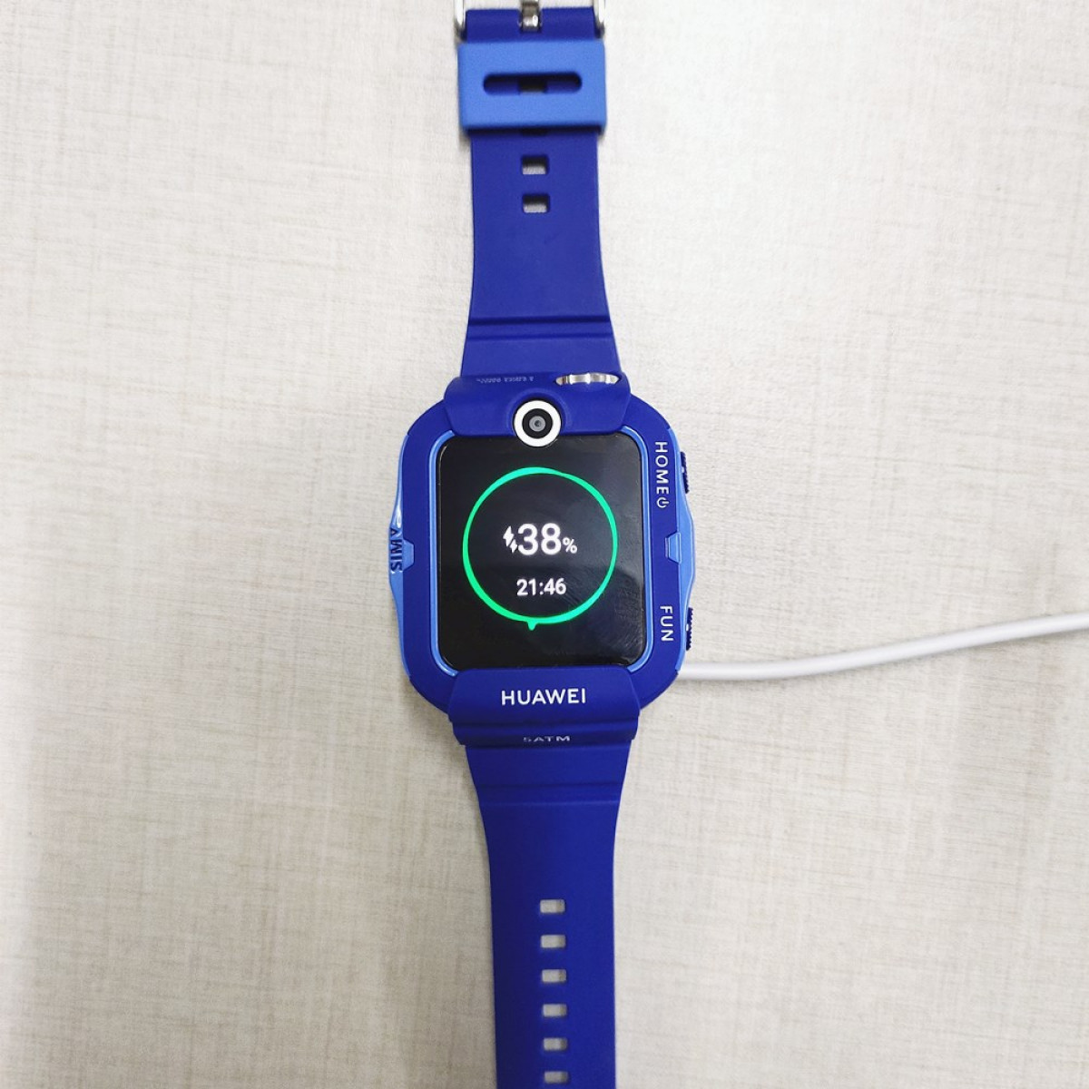 Niebieski smartwatch wyświetlający godzinę, procent naładowania baterii i status ładowania na drewnianej powierzchni.