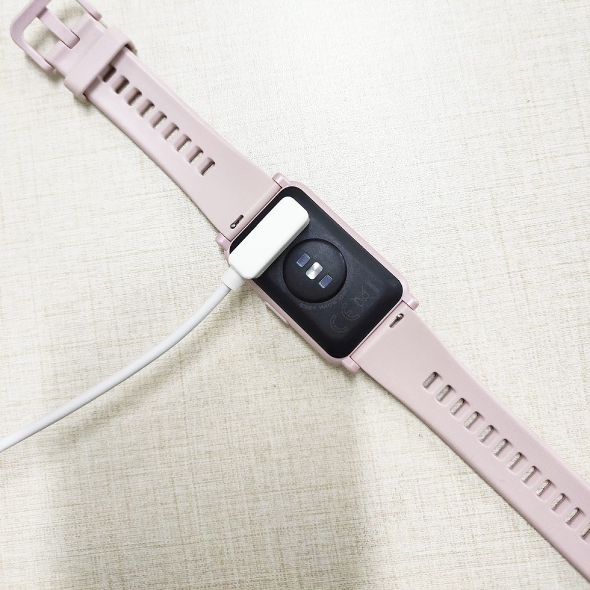 Różowy smartwatch z podłączonym białym kablem do ładowania. Zegarek leży na powierzchni.