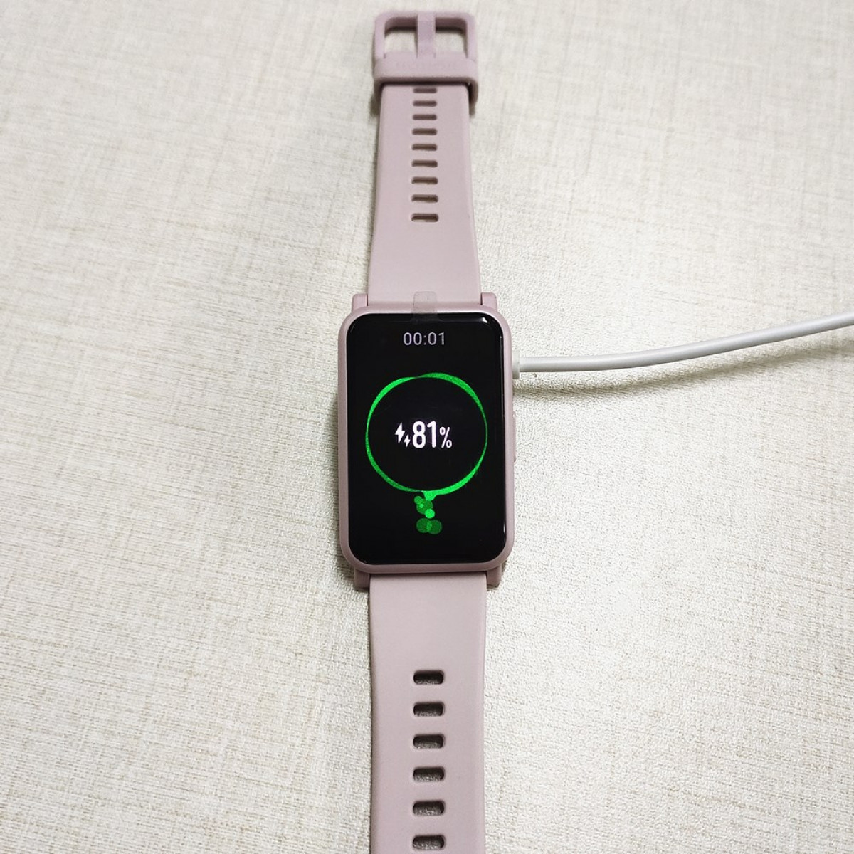 Różowy smartwatch w trakcie ładowania, pokazujący 48% naładowania. Kremowa powierzchnia. Biały kabel ładujący.