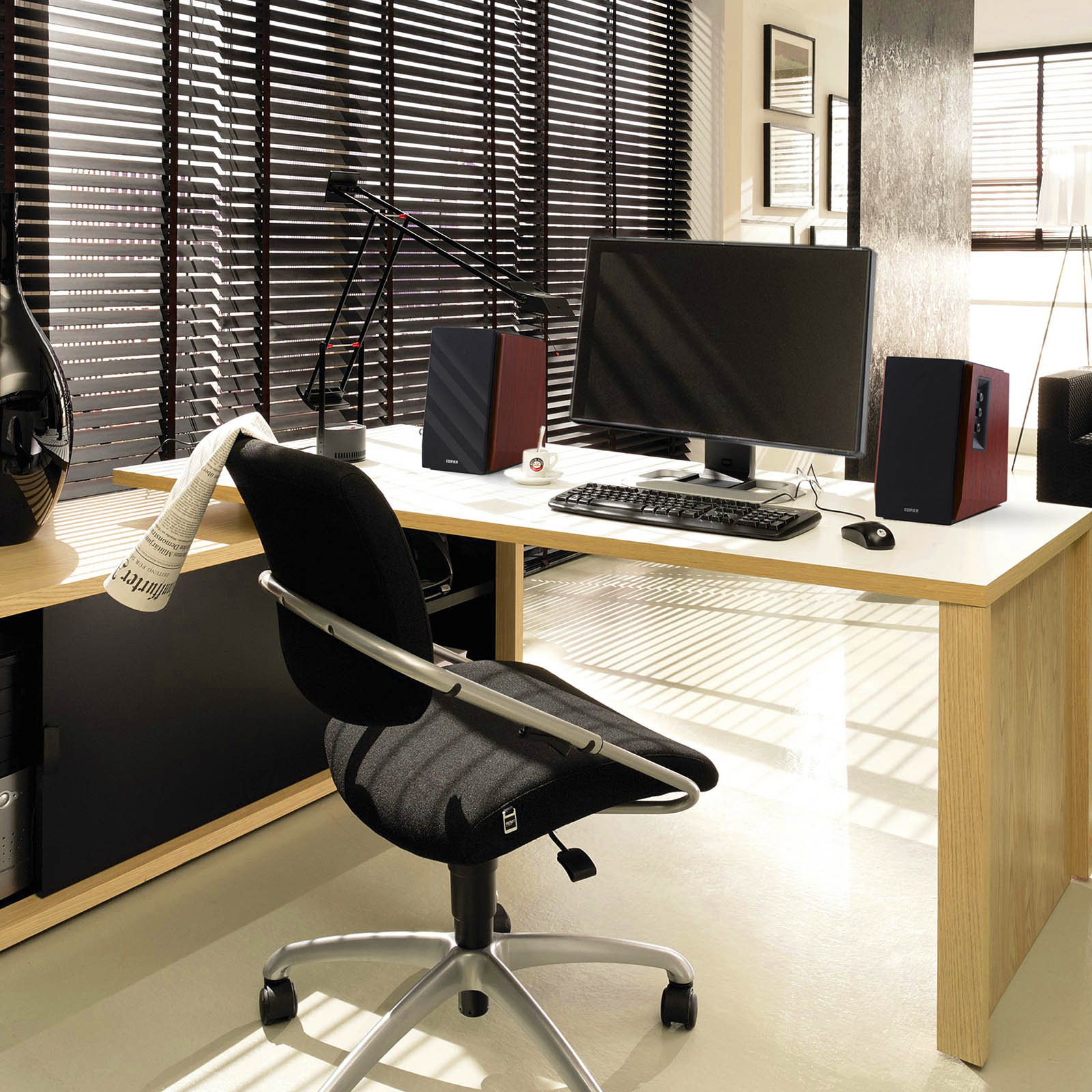 Biuro z biurkiem, komputerem, głośnikami, krzesłem i żaluzjami. Filiżanka i gazeta.