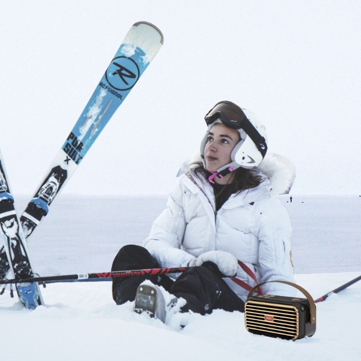 Frau in Schneeausrüstung sitzt in der Nähe von Skiern und einem Lautsprecher im Schnee.