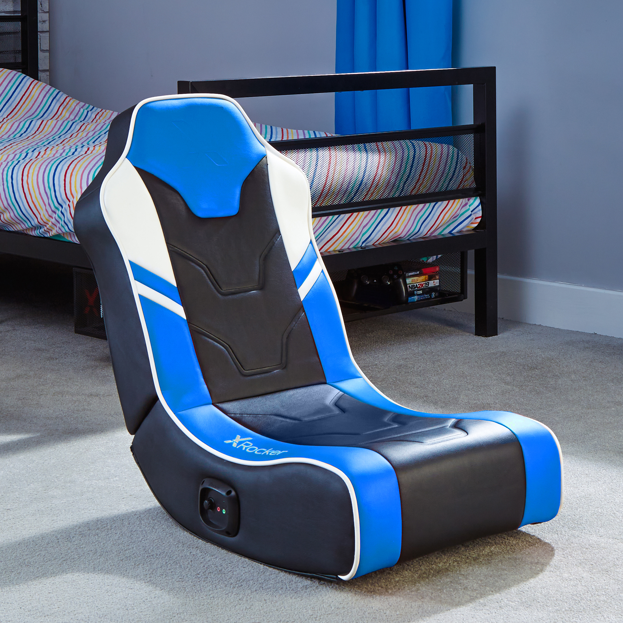 Gaming-Stuhl, blau und schwarz. Zeigt den Stuhl in einem Raum mit Bett, Teppich und Regal mit Spielen.