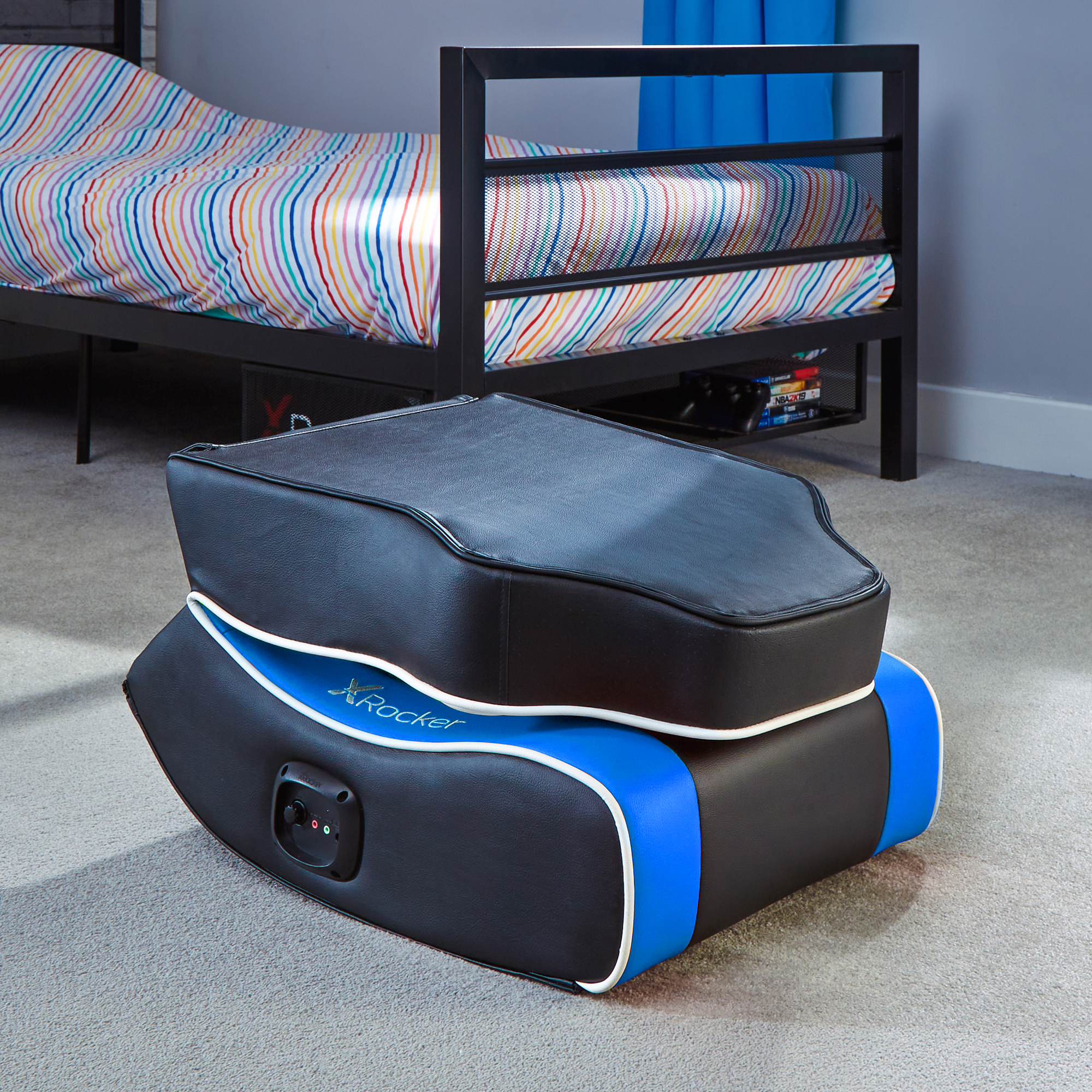 Ein schwarzer und blauer Gaming-Stuhl steht auf einem Teppichboden. Dahinter steht ein Bett mit einer gestreiften Bettdecke.