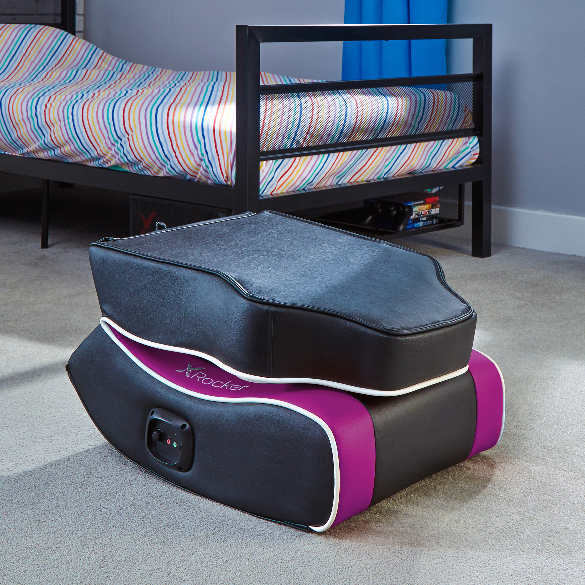 Gaming-Stuhl auf einem Teppich. Der Stuhl ist schwarz, lila und weiß. Bett und Schreibtisch im Hintergrund.