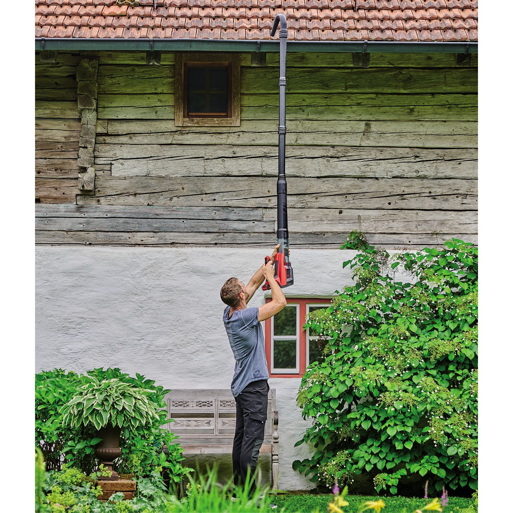 Mężczyzna używa czarno-czerwonego urządzenia do czyszczenia rynien. W tle drewniany dom.